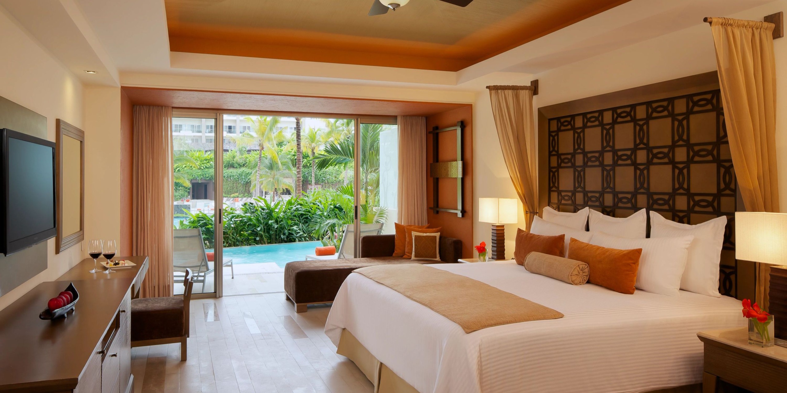 The Dreams Vallarta Bay features a bedroom with an expansive bed, elegantly decorated in brown and beige tones, including a comfortable sofa and a sleek flat-screen TV. Sliding glass doors lead out to a patio that offers stunning views of the pool and garden.
