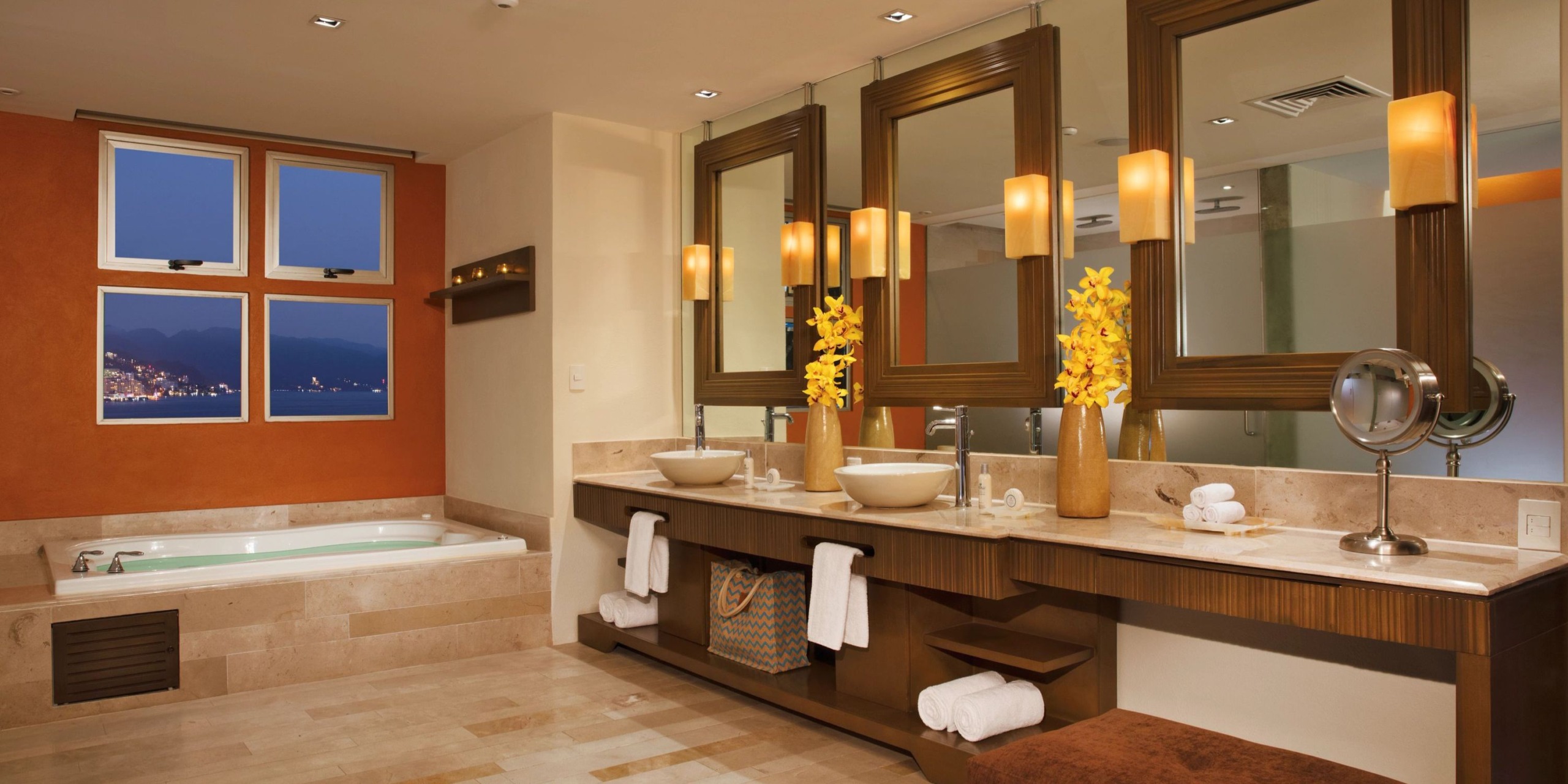 Transform your bathroom into a luxurious oasis with the Dreams Vallarta Bay double vanity featuring two elegant vessel sinks and large mirrors. Its wooden accents perfectly complement the spa bathtub, creating a serene atmosphere. Enhancing the elegance are warm lighting and vibrant yellow flowers, all while offering stunning cityscape views through the window.