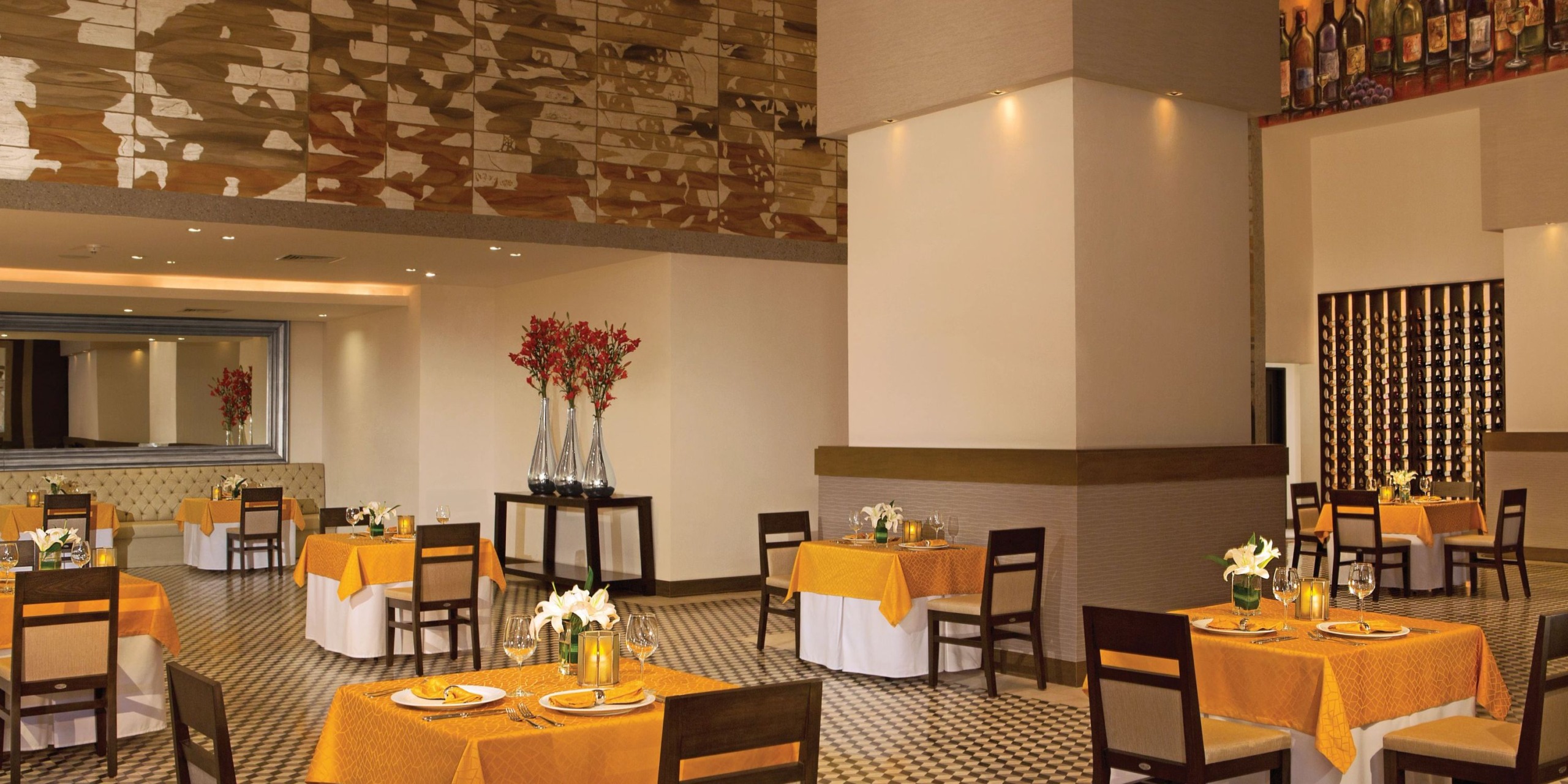 Dreams Vallarta Bay features a dining area adorned with yellow tablecloths, dark wood chairs, and vases filled with flowers. The walls are decorated with artworks and a wine rack, while the floor boasts a geometric pattern. Elegantly set tables showcase candles and glassware.