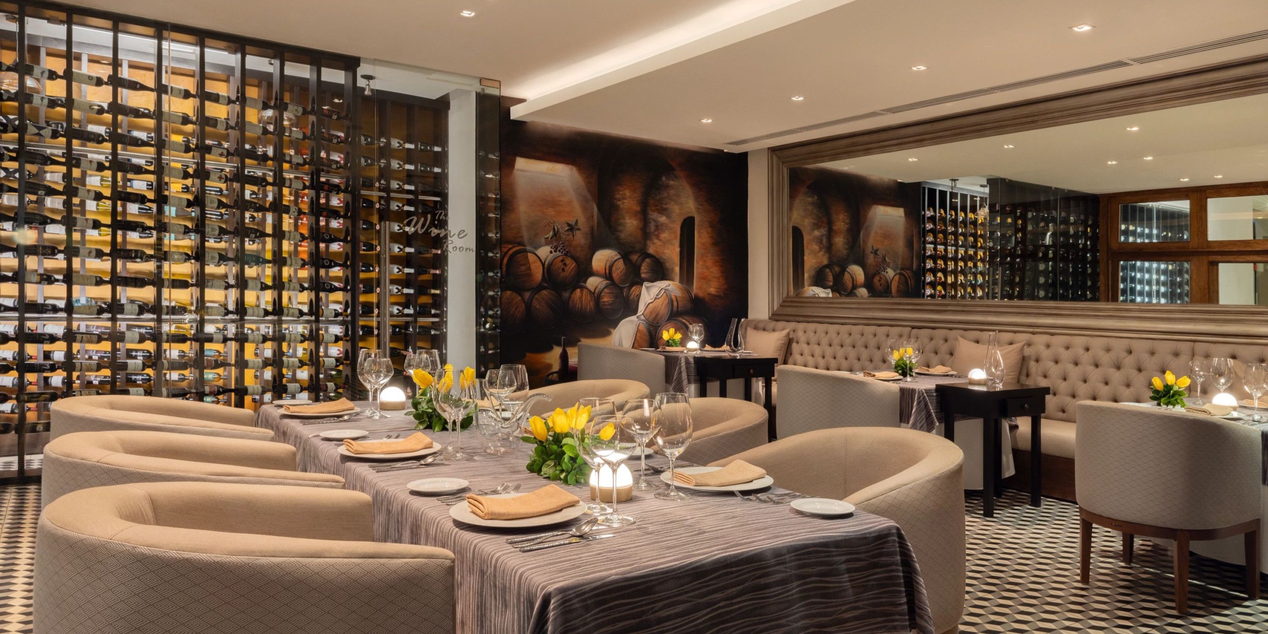 The Dreams Vallarta Bay offers an elegant interior with soft beige seating, a wall of wine bottles, and tables set with yellow flowers and glasses. Dim lighting enhances the cozy atmosphere.