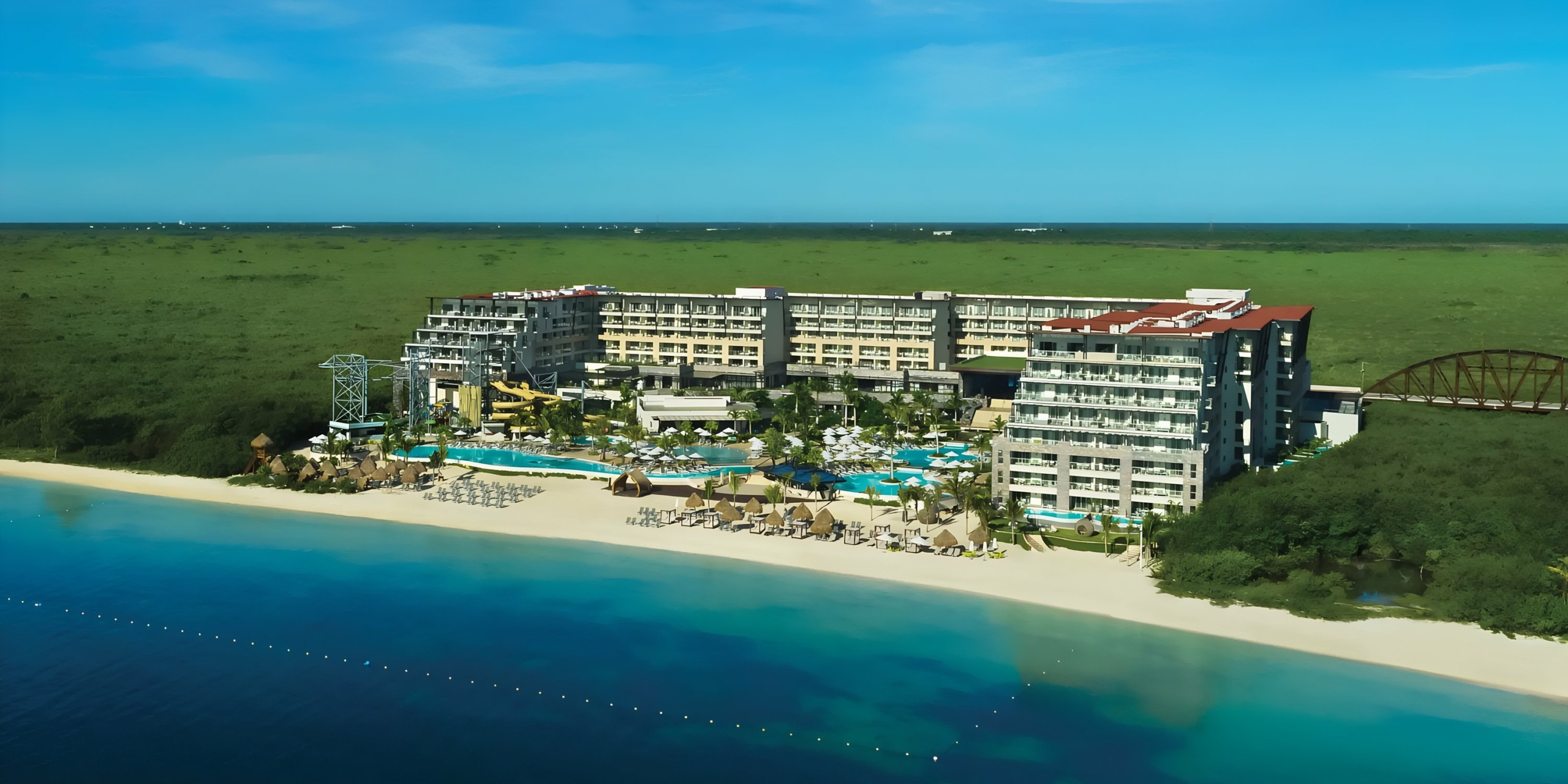 A Dreams Natura resort with swimming pools and sun loungers, adjacent to a beach and overlooking a vast green landscape.