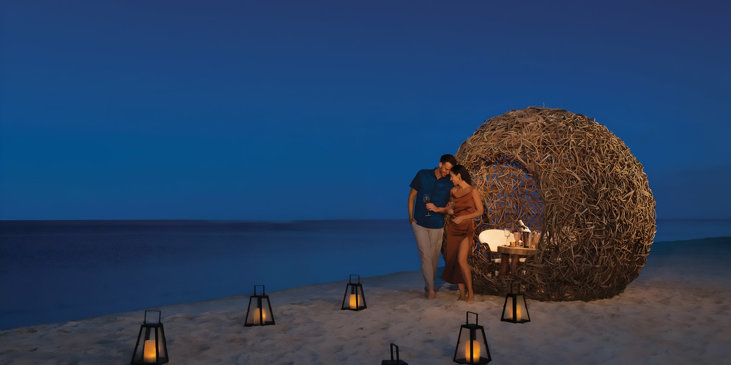 A couple enjoys a romantic evening on the beach with a dinner setup by Dreams Natura and lanterns, under a dusky sky.