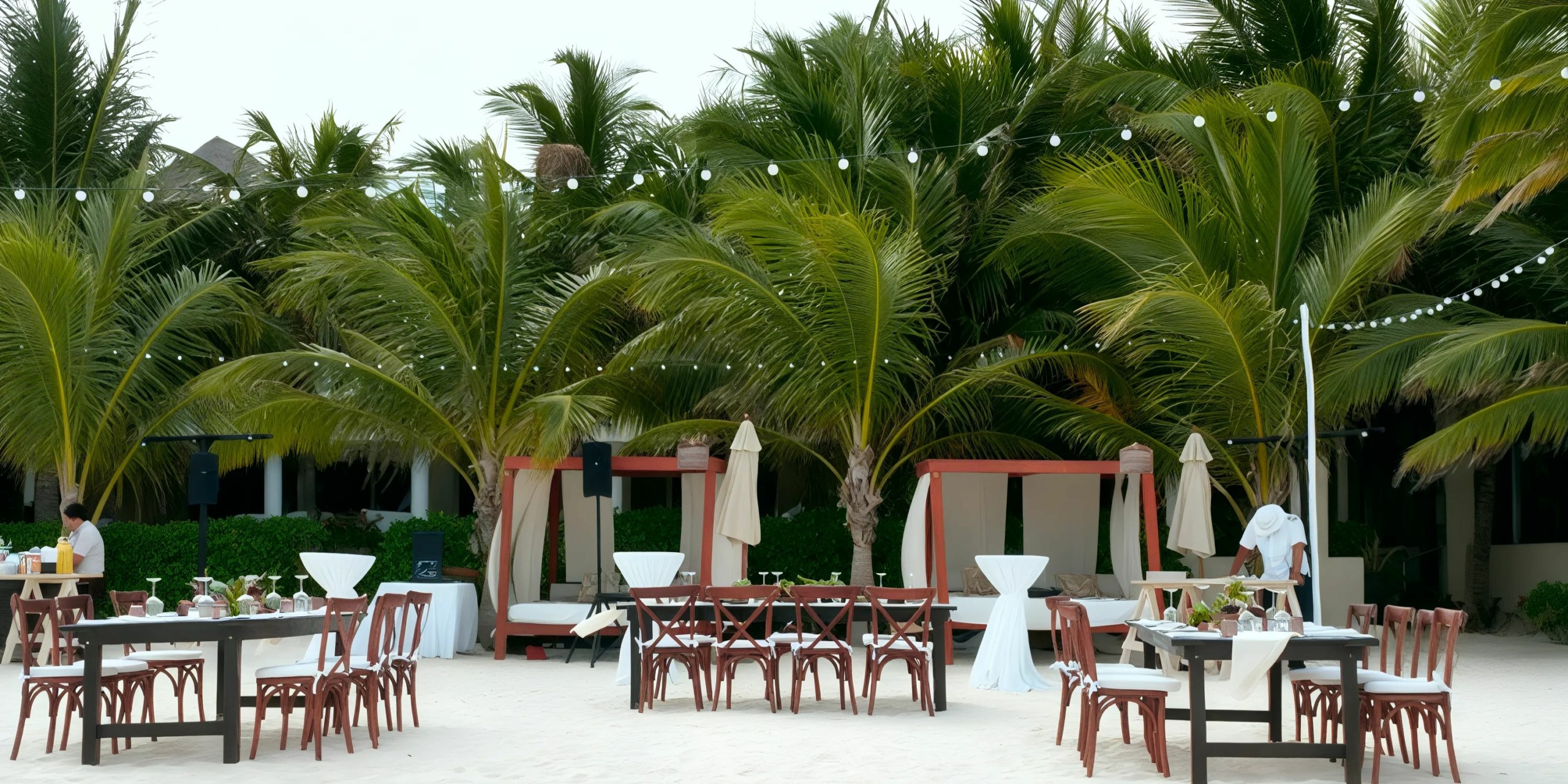 Outdoor beach dining setup with decorated tables and chairs, surrounded by palm trees. String lights are hanging above, creating a festive atmosphere.
