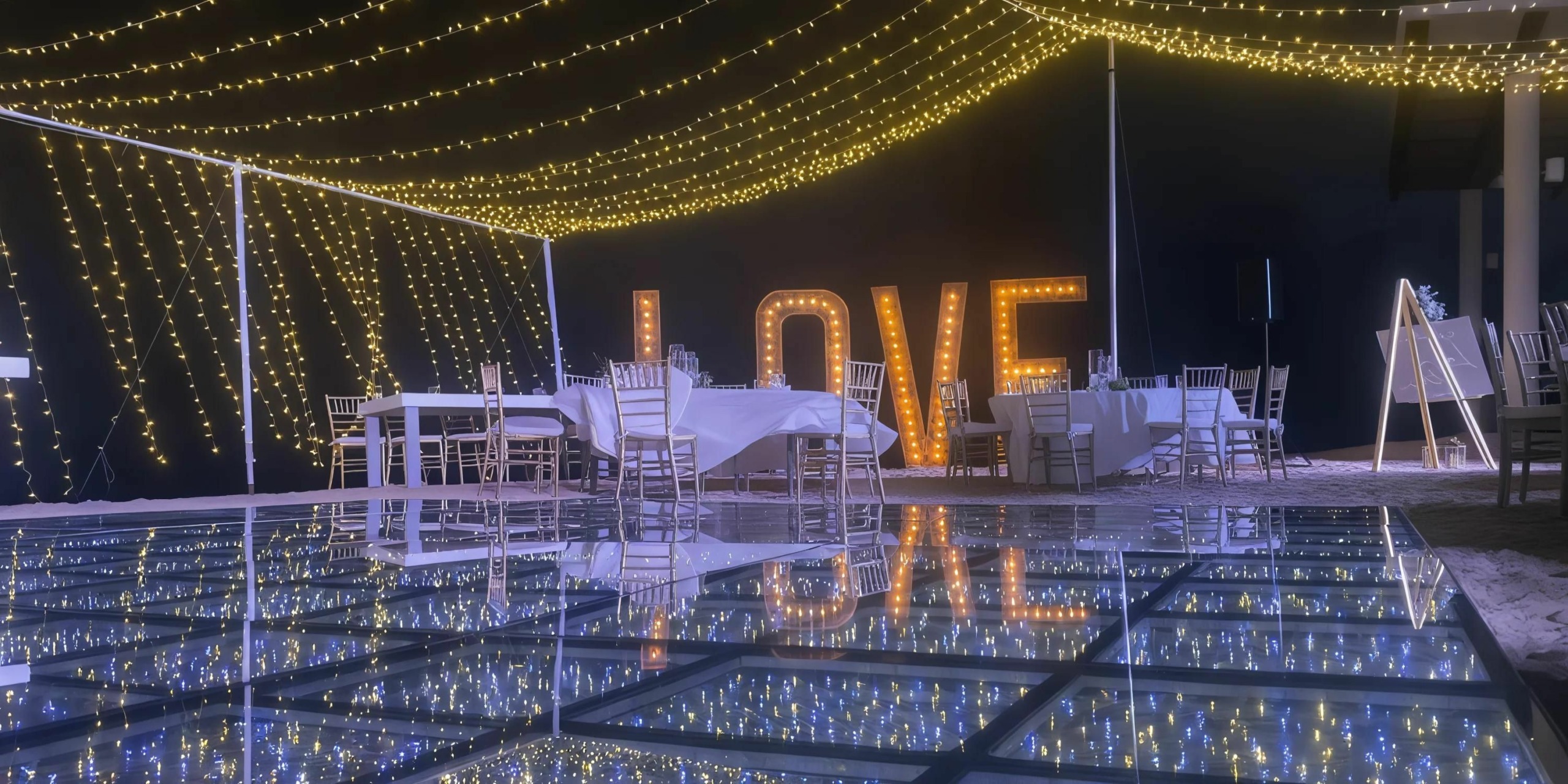 A wedding reception setup at the Breathless Riviera Cancún Resort and Spa features tables, chairs, and string lights. The word "LOVE" is illuminated in large letters, and there is a reflective glass floor.
