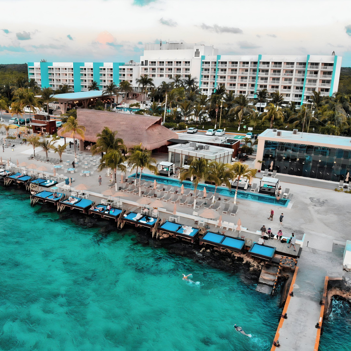 Aerial view of a Fiesta Americana Cozumel destination wedding at a tropical resort with beachfront cabanas, clear blue waters, and a large hotel building surrounded by palm trees during sunset.