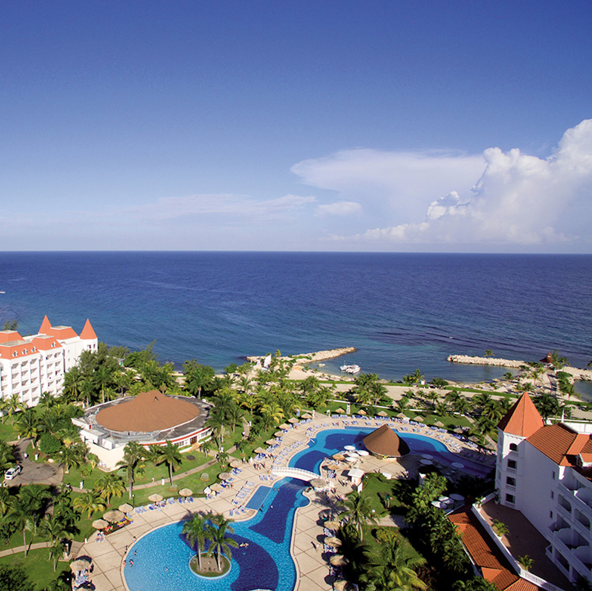 Aerial view of Bahia Principe Luxury Runaway Bay, a perfect destination wedding venue with a large swimming pool, white buildings, and a clear view of the ocean under a blue sky.