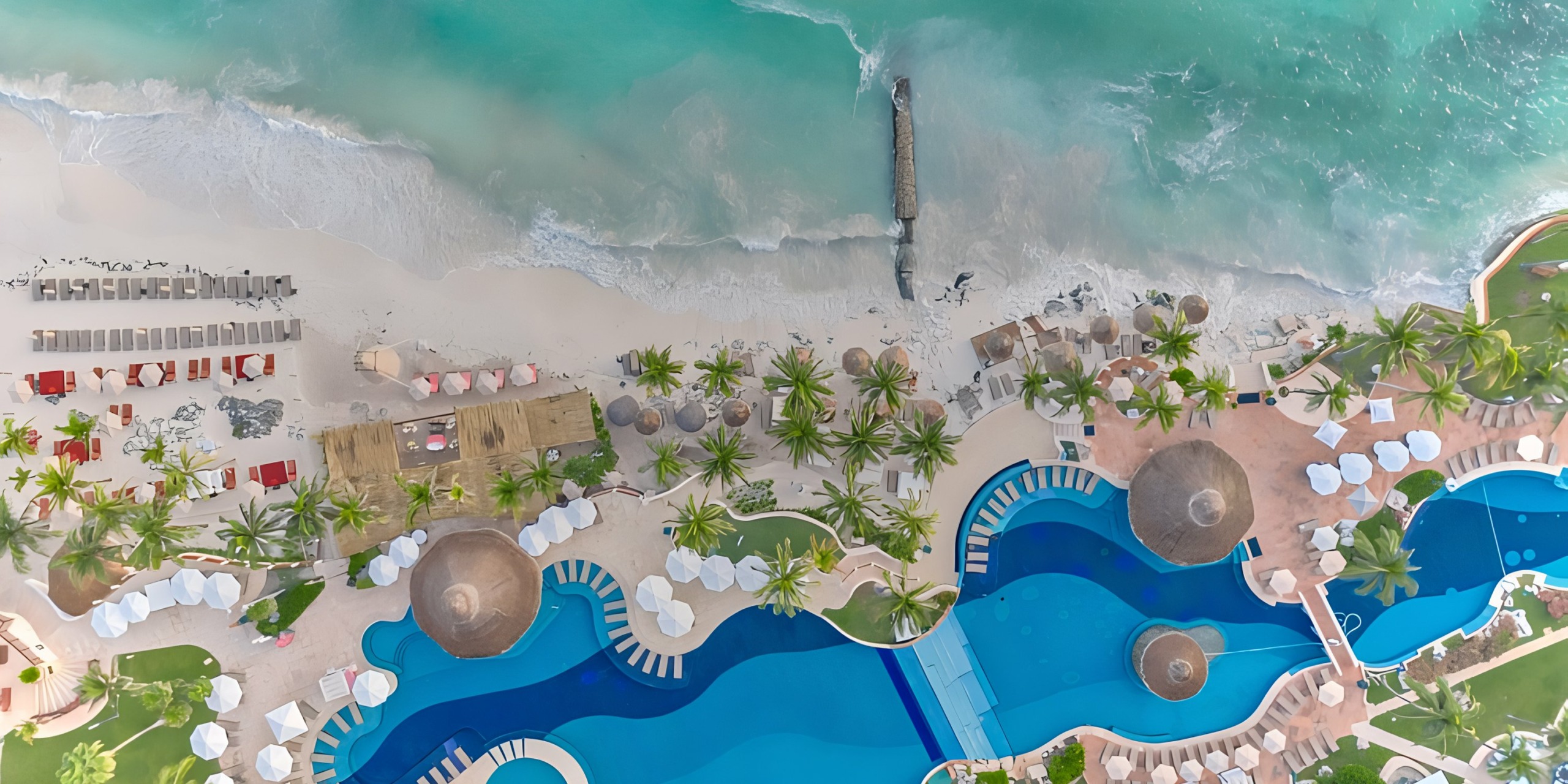 Aerial view of Grand Fiesta Americana Coral Beach resort with blue swimming pools, palm trees, white sandy beach, and turquoise ocean waves.