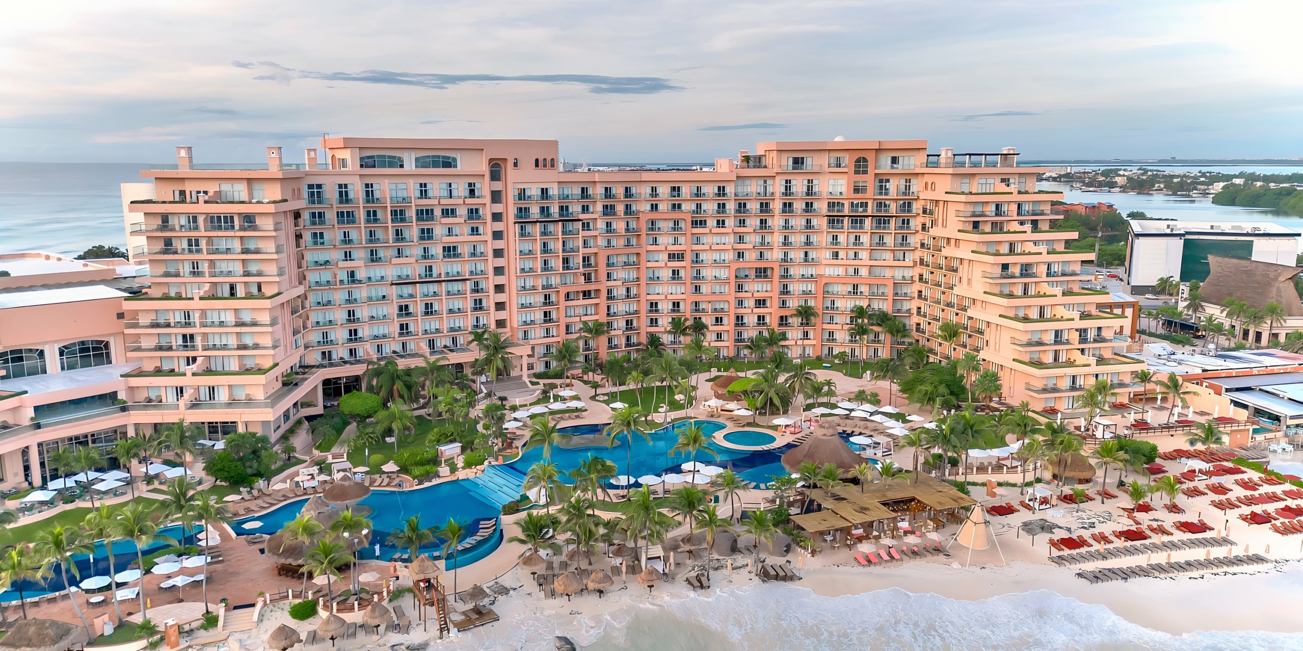 Aerial view of the Grand Fiesta Americana Coral Beach, a large beachfront hotel with multiple swimming pools, surrounded by palm trees, during early evening.