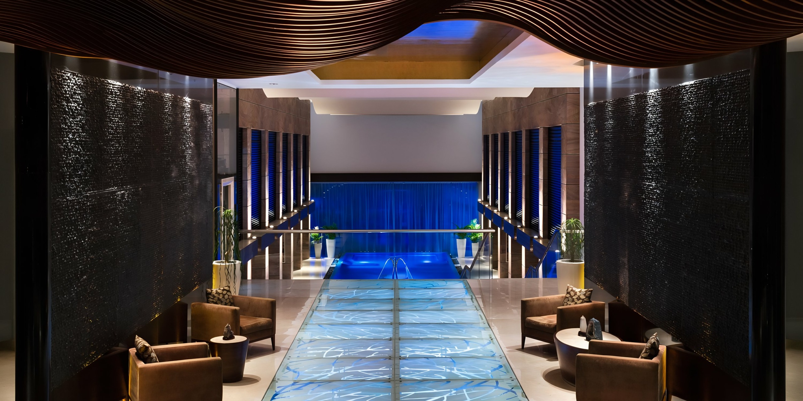 A luxurious Grand Fiesta Americana Coral Beach hotel lobby with a central blue-lit walkway, flanked by dark water features and warm ambient lighting from above.