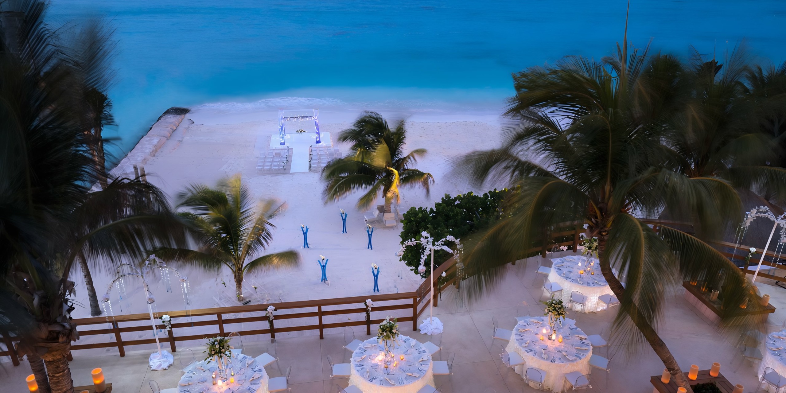 Aerial view of a Grand Fiesta Americana Coral Beach evening event with tables set for a dinner, palm trees, and a lighted gazebo by the turquoise sea.
