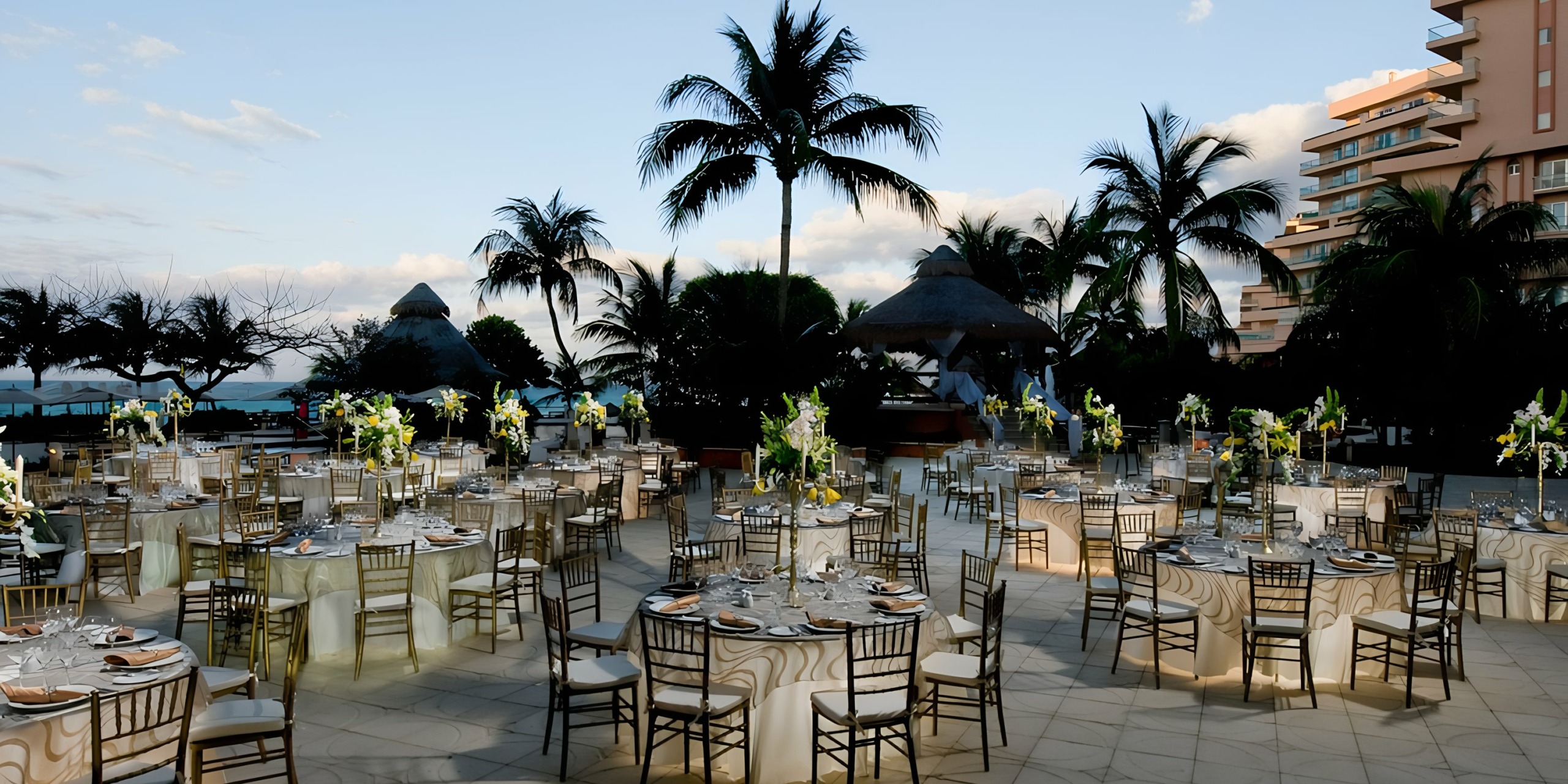 Outdoor wedding reception setup at twilight with round tables, floral centerpieces, and palm trees near the Grand Fiesta Americana Coral Beach coastal backdrop.