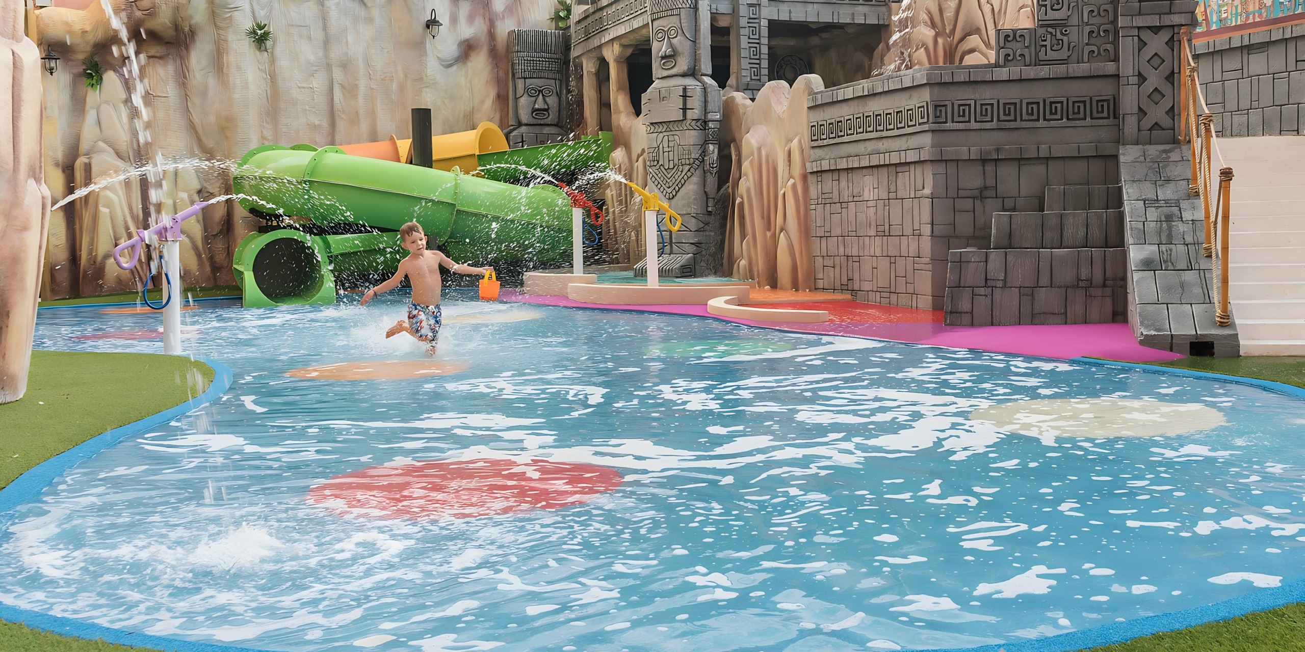 A child runs through the Grand Fiesta Americana Coral Beach with sprays of water, surrounded by themed ruins and slides.