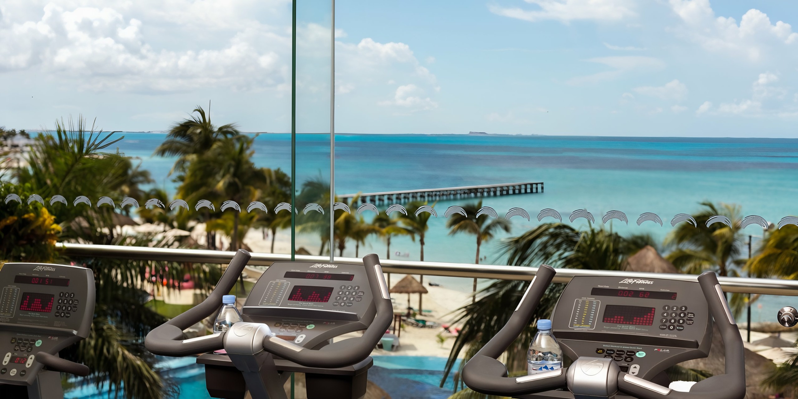 View from the Grand Fiesta Americana Coral Beach gym treadmill overlooking a tropical beach with clear blue skies, palm trees, and a turquoise ocean.