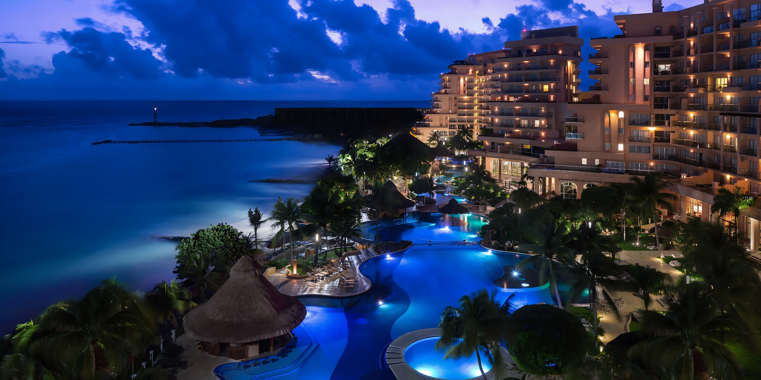 Nighttime view of Grand Fiesta Americana Coral Beach and pool beside the ocean, with lights reflecting on water and a distant lighthouse visible.