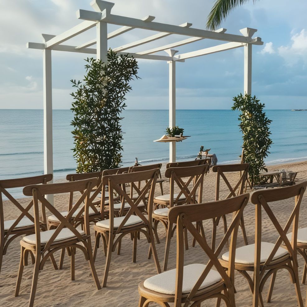 Chairs are set up on a sandy beach, all oriented towards a white arch adorned with lush greenery, creating the perfect setting for a destination wedding beside the tranquil sea.