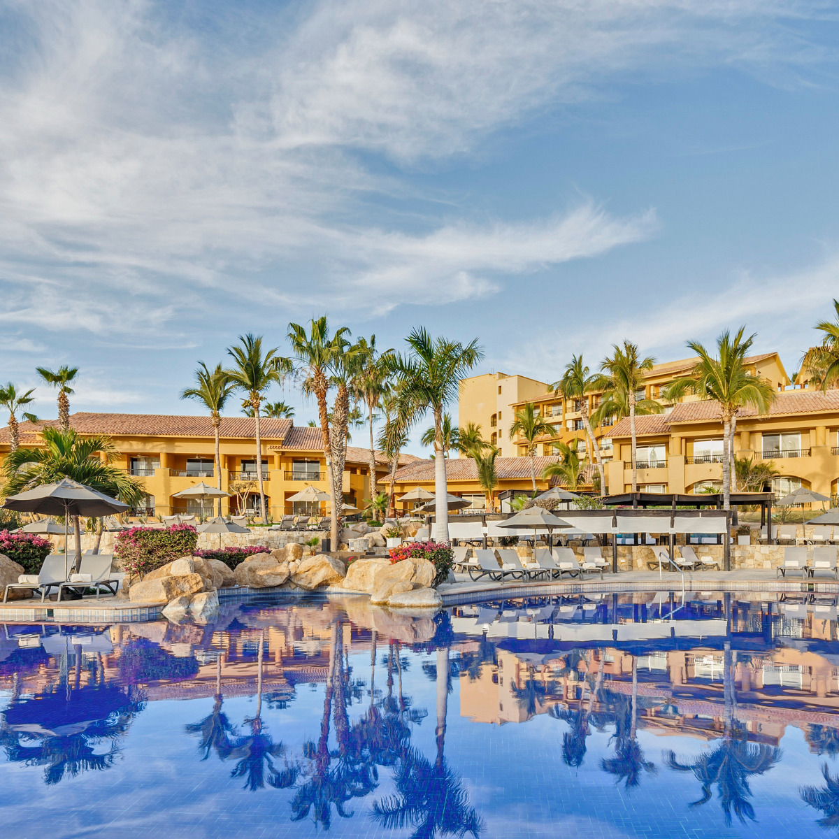 A Grand Fiesta Americana Los Cabos, perfect for a destination wedding, with palm trees and lounge chairs reflected in a clear blue swimming pool at sunrise.