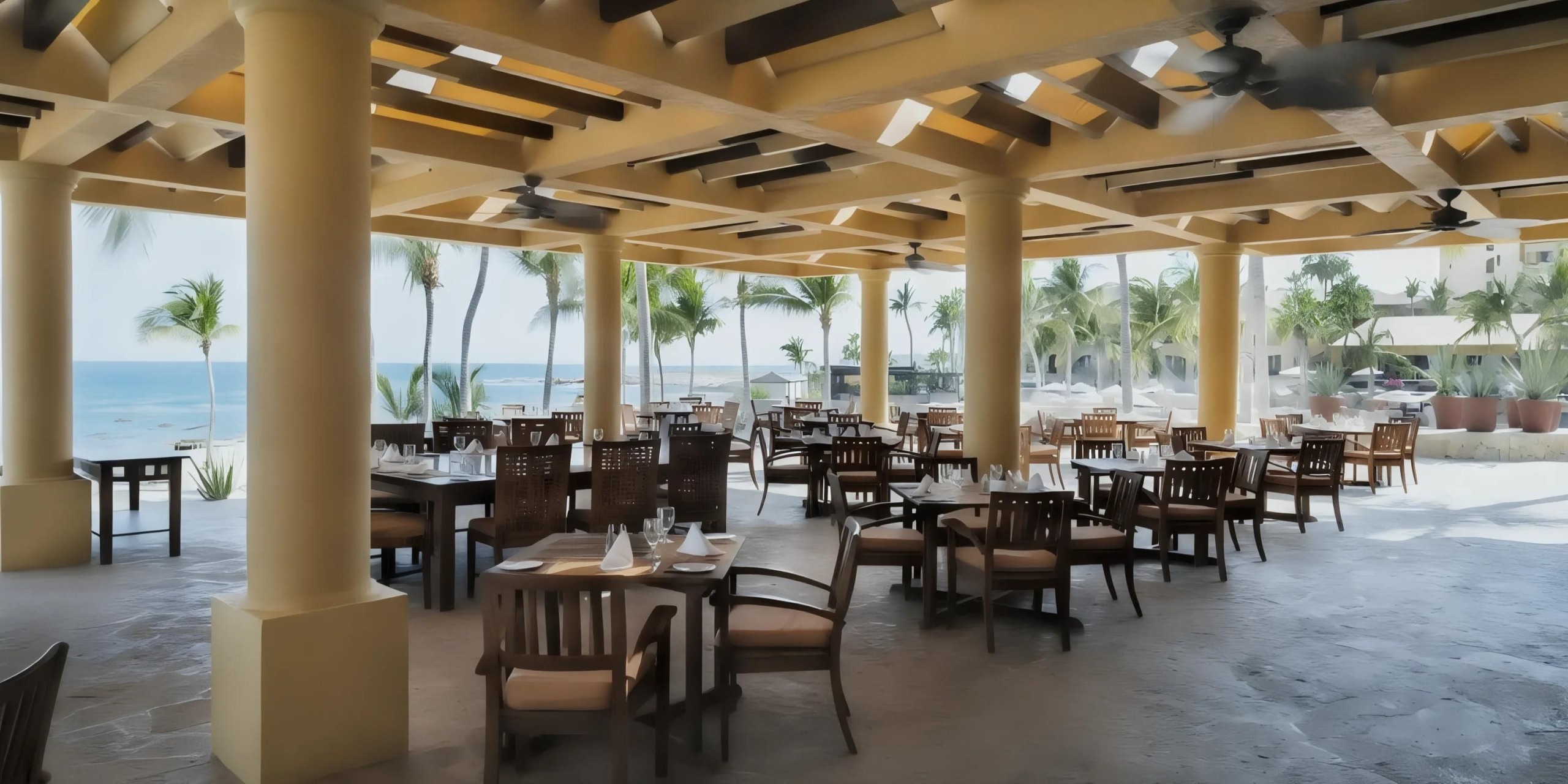 Outdoor restaurant with wooden tables and chairs under a pergola, overlooking a beach with palm trees and ocean views.