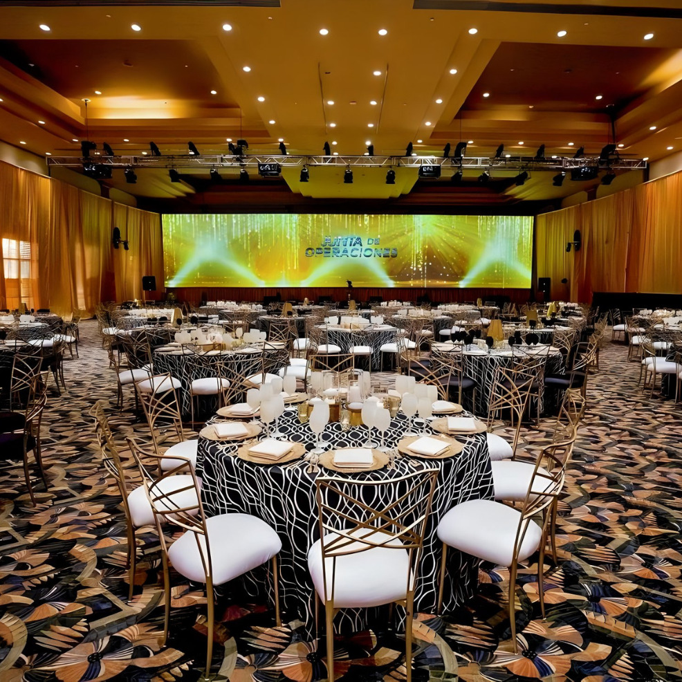 Elegant banquet hall setup for a destination wedding, featuring round tables with white chairs, stylish table settings, and a large screen displaying vibrant green graphics at the front.