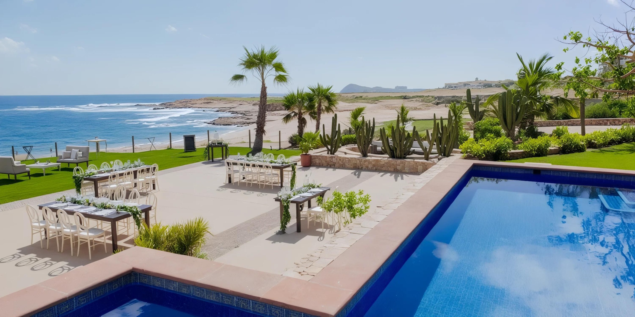 A seaside patio with tables, chairs, and a pool, overlooking the ocean and a sandy beach. Palm trees and cacti adorn the landscape.