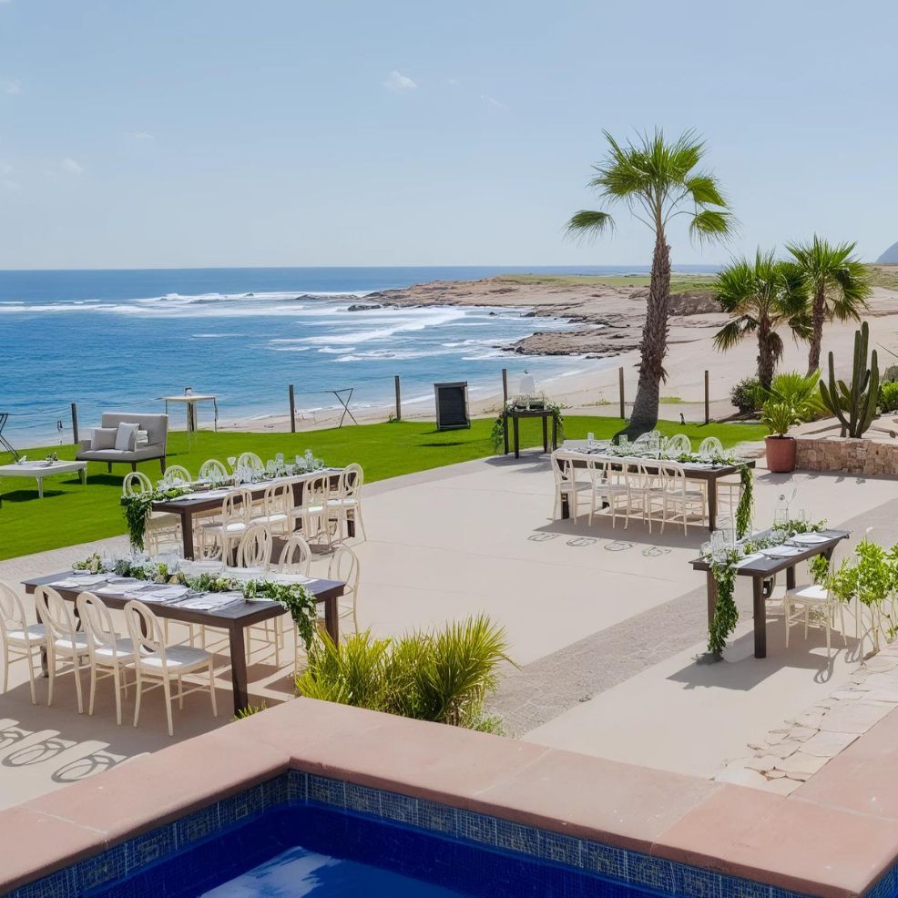 Outdoor event setup by the ocean with tables and chairs on a patio, surrounded by greenery and palm trees, overlooking a sandy beach.