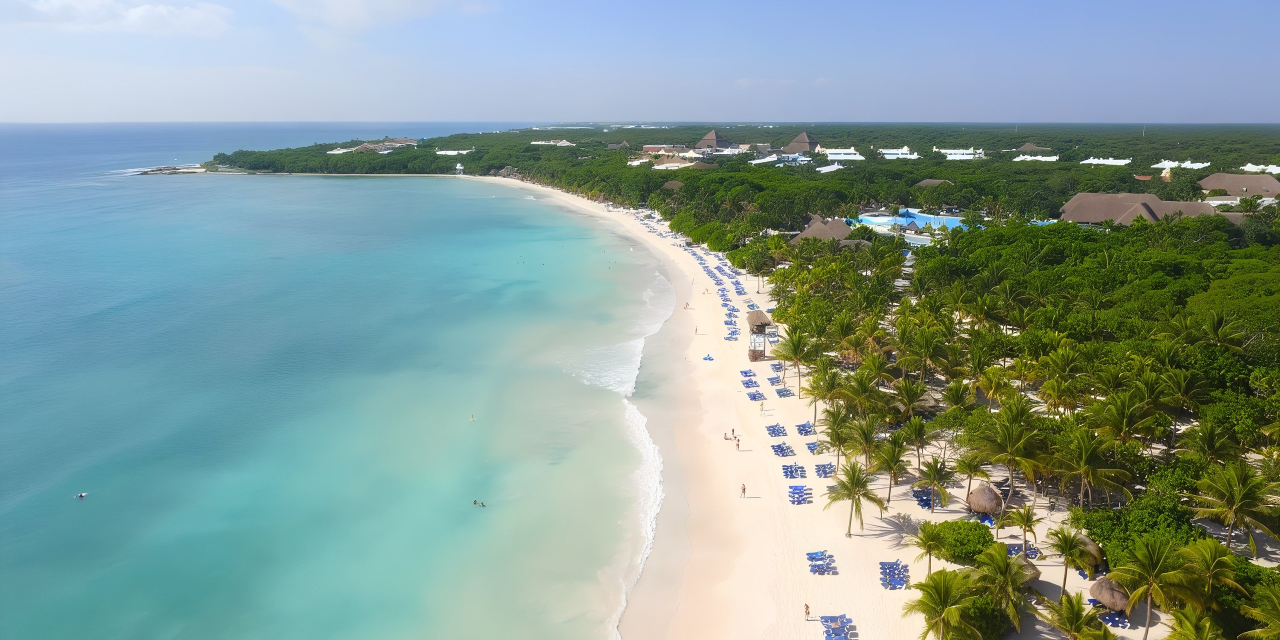 Experience a breathtaking aerial view of the Grand Palladium Kantenah, featuring a tropical beach with pristine white sand, crystal-clear turquoise waters, and lush greenery. Relaxing beach chairs and umbrellas line the picturesque shoreline, with captivating buildings visible in the background.