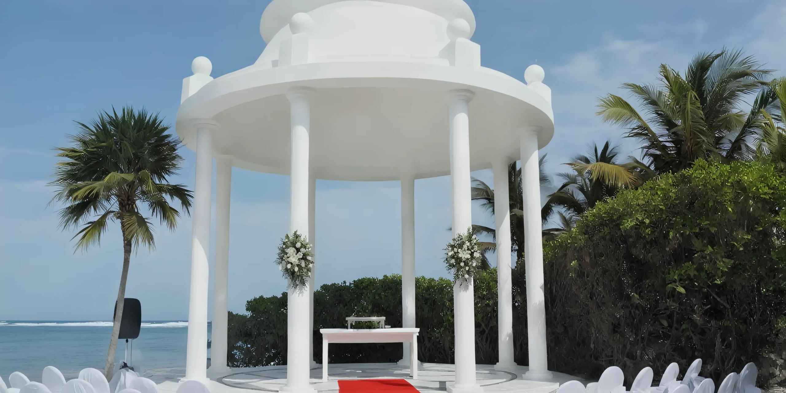 A round, white gazebo with columns stands on a red carpet setup for an event near the sea, with rows of white chairs facing it. Palm trees and lush greenery surround the area.