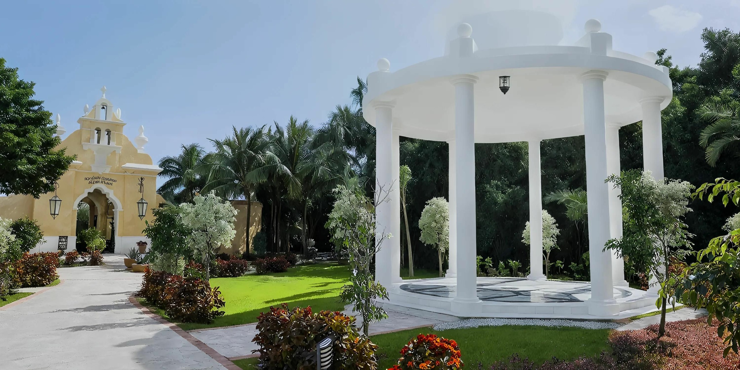 A white gazebo with columns stands near a landscaped garden path leading to a yellow building with an arched entrance, surrounded by lush greenery and flowering plants under a clear blue sky.