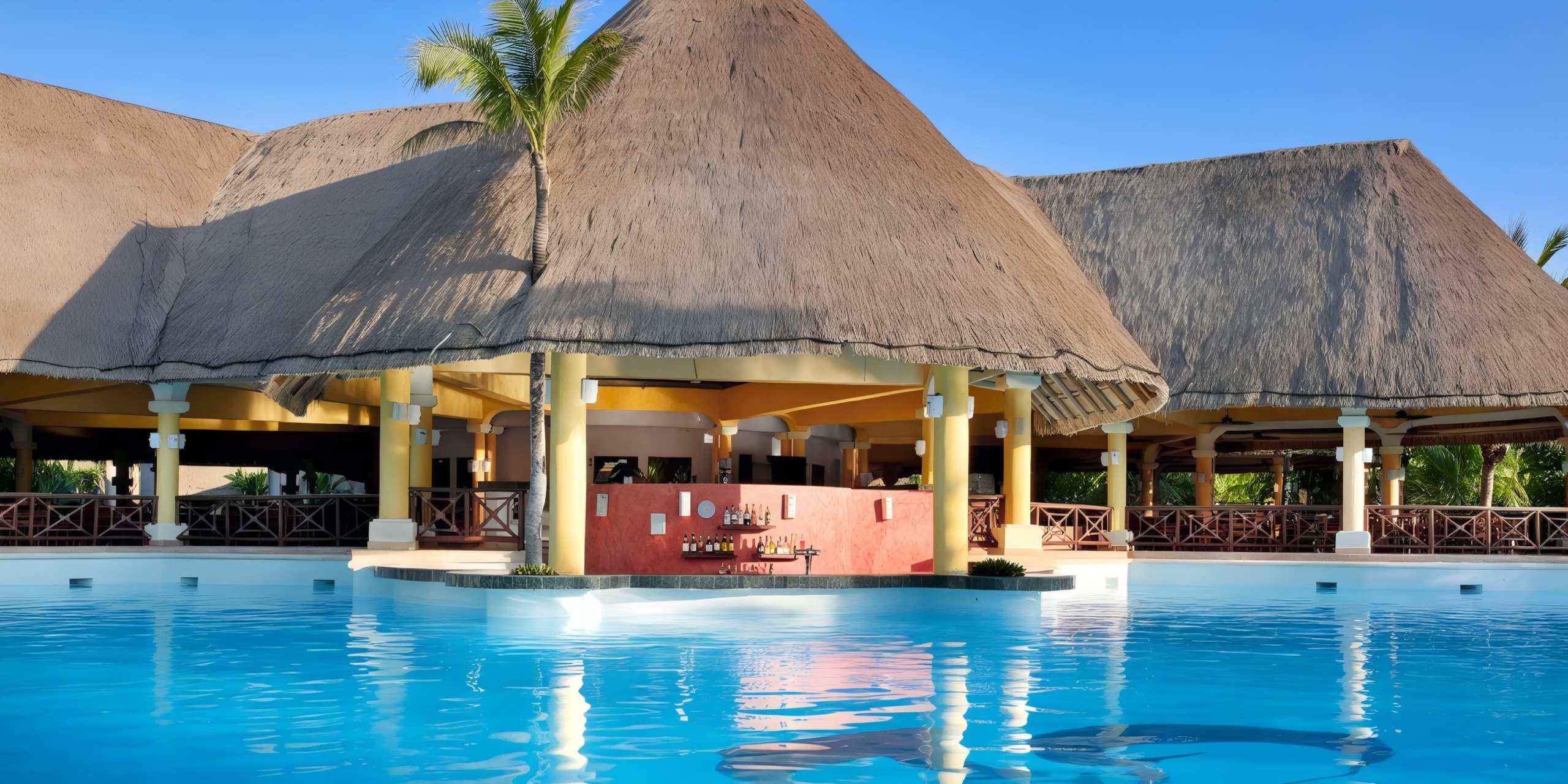 In front of the thatched-roof bar, surrounded by columns and lush greenery under a clear blue sky, lies the expansive outdoor swimming pool at Grand Palladium Kantenah.