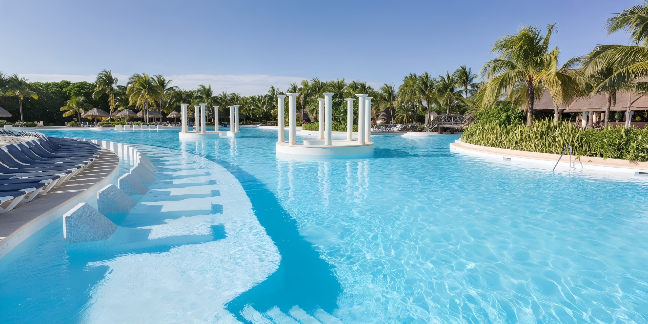 The Grand Palladium Kantenah features a spacious outdoor swimming pool with pristine blue water, encircled by palm trees and sun loungers. Floating columns grace the center, while huts are nestled in the background beneath a clear sky.