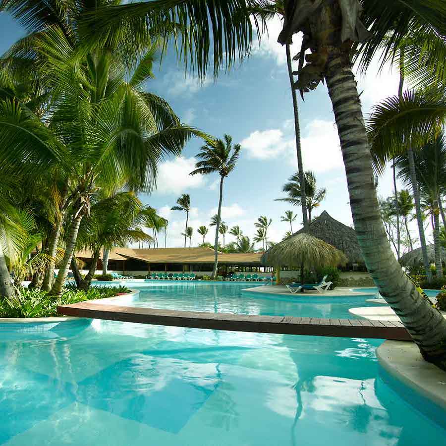 Grand Palladium Punta Cana destination wedding resort swimming pool surrounded by palm trees with a thatched roof pavilion under a clear blue sky.