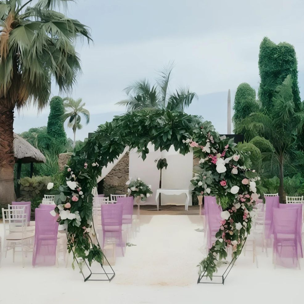 Outdoor wedding setup with a floral arch, pink-draped chairs, and a small altar in a tropical garden setting.