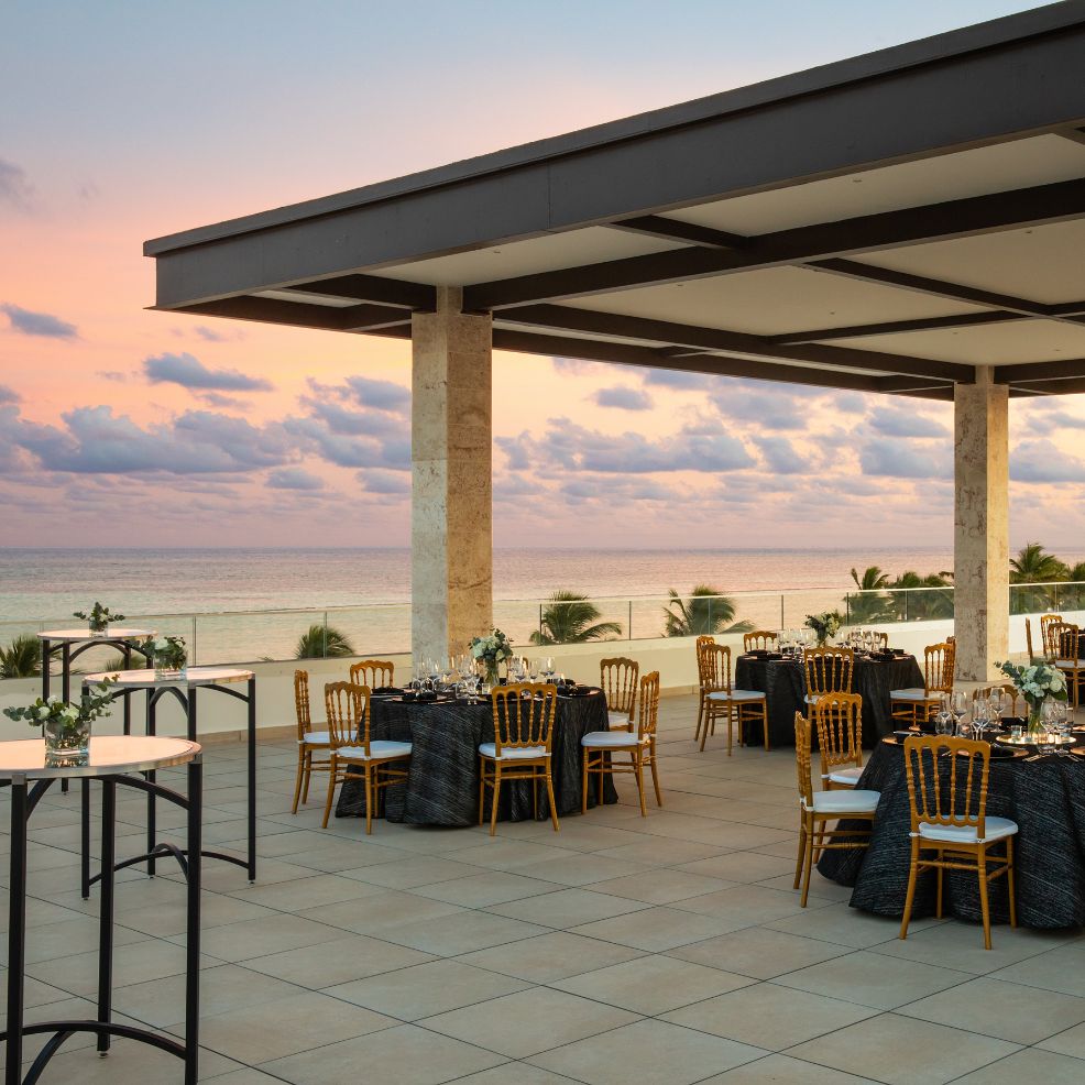 An outdoor terrace equipped with dining tables and chairs provides a breathtaking view of the ocean at sunset, creating the perfect setting for a romantic destination wedding.