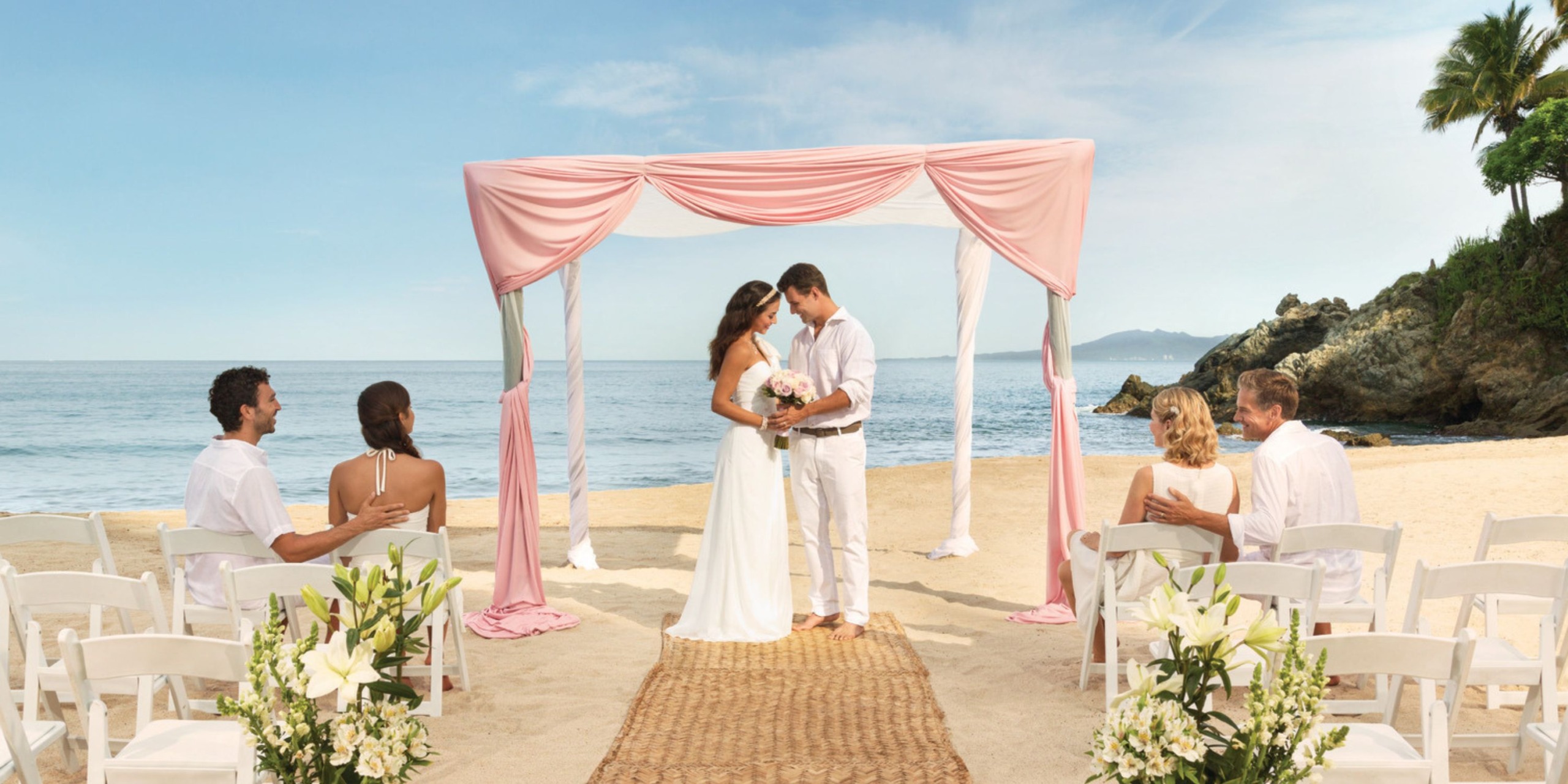 A destination wedding at Hyatt Ziva features a couple standing beneath a pink arch on the beach, surrounded by their seated guests.