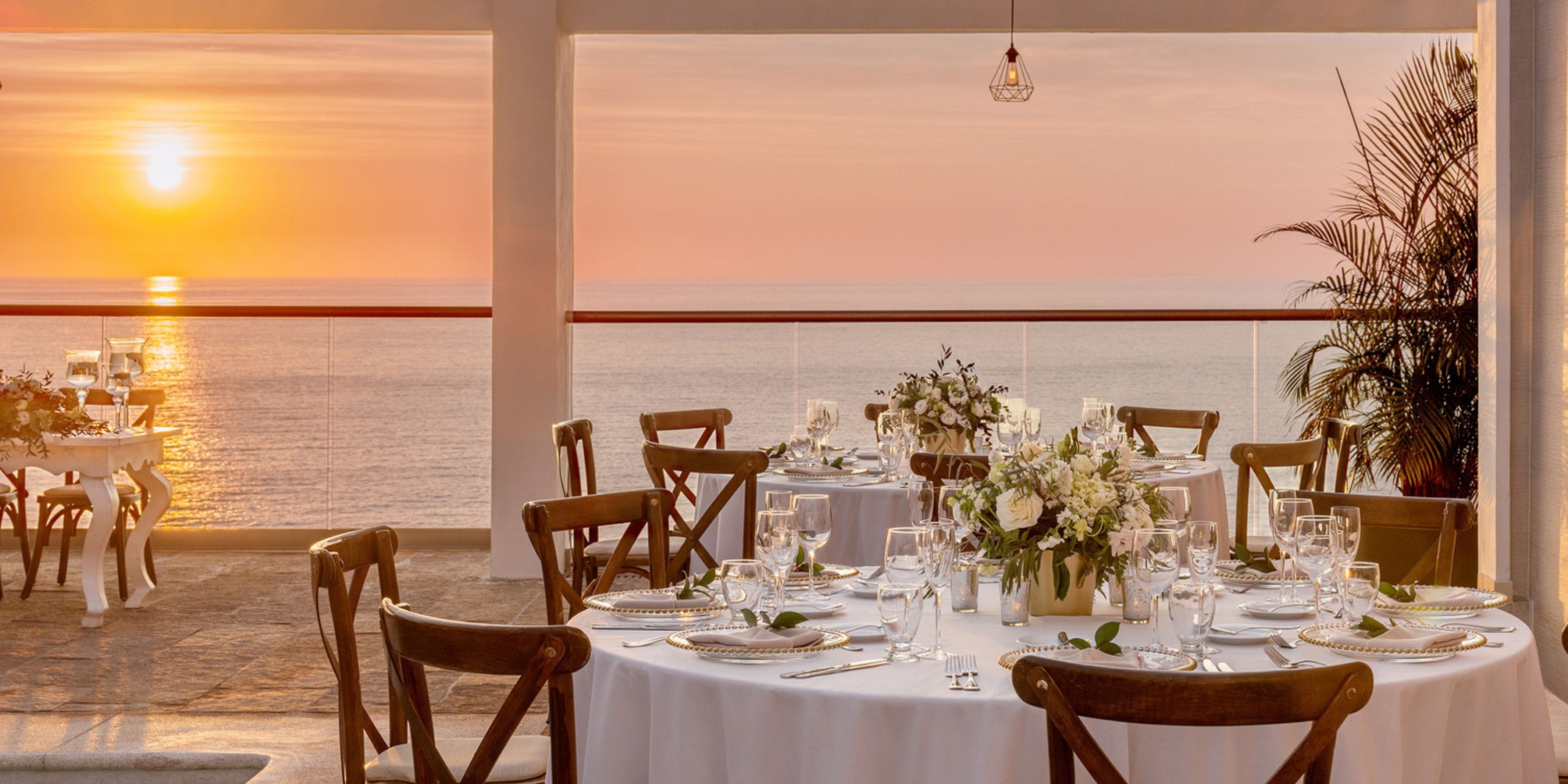 A breathtaking sunset view enhances an elegant dining setup adorned with white tablecloths and floral centerpieces, perfect for a dreamy destination wedding.