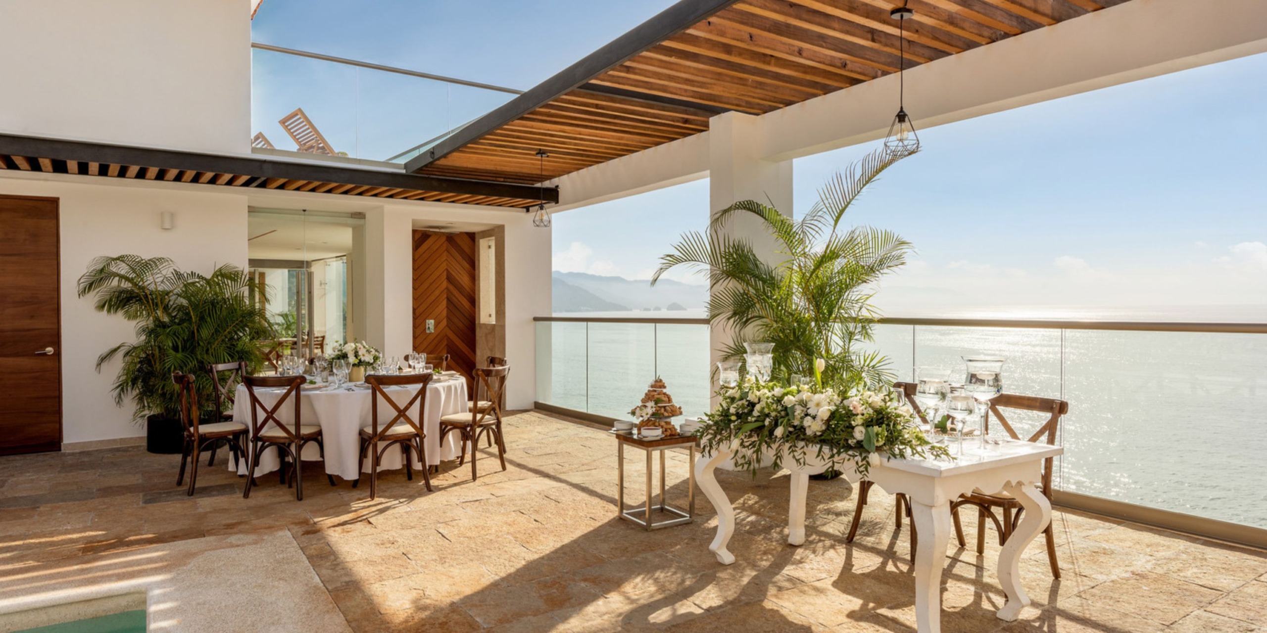 A rooftop patio beautifully arranged for a destination wedding, featuring two elegantly set tables, vibrant floral arrangements, and a breathtaking ocean view.
