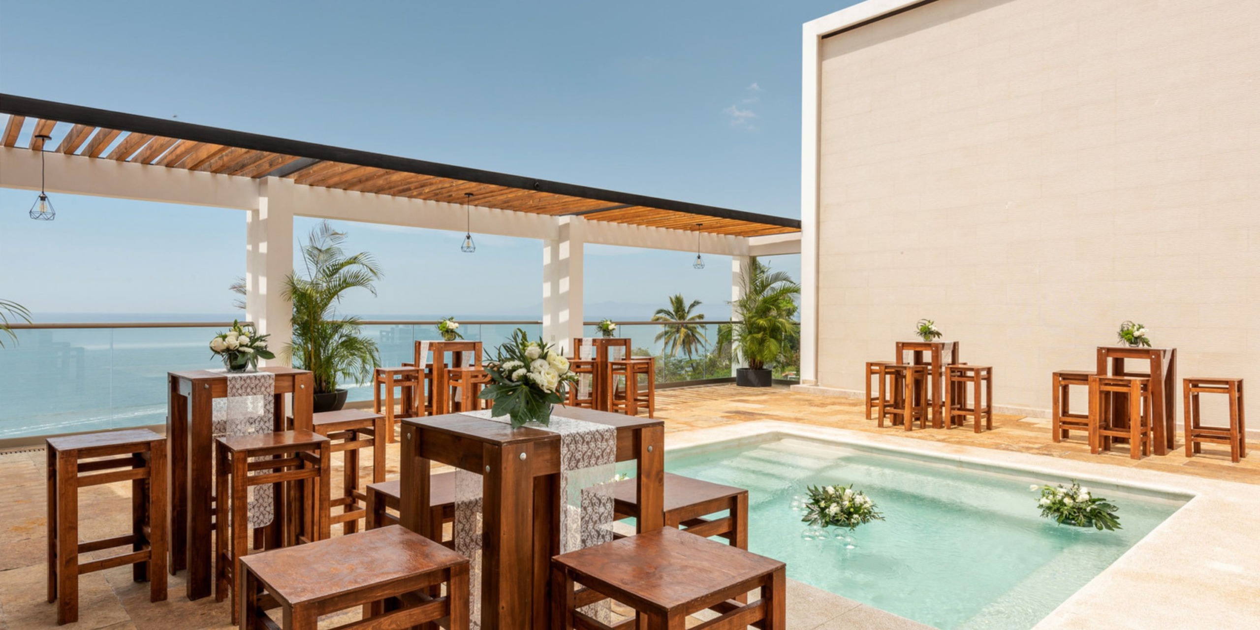 Experience a destination wedding on an outdoor terrace featuring wooden tables, a pool, and stunning ocean views beneath a blue sky.