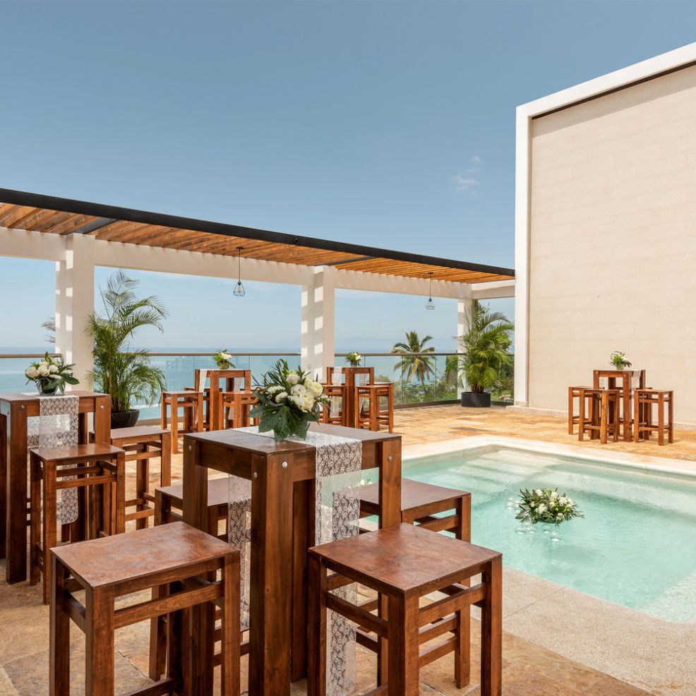 Outdoor poolside area featuring tall wooden tables and chairs, with a breathtaking ocean view in the background—perfect for a destination wedding.