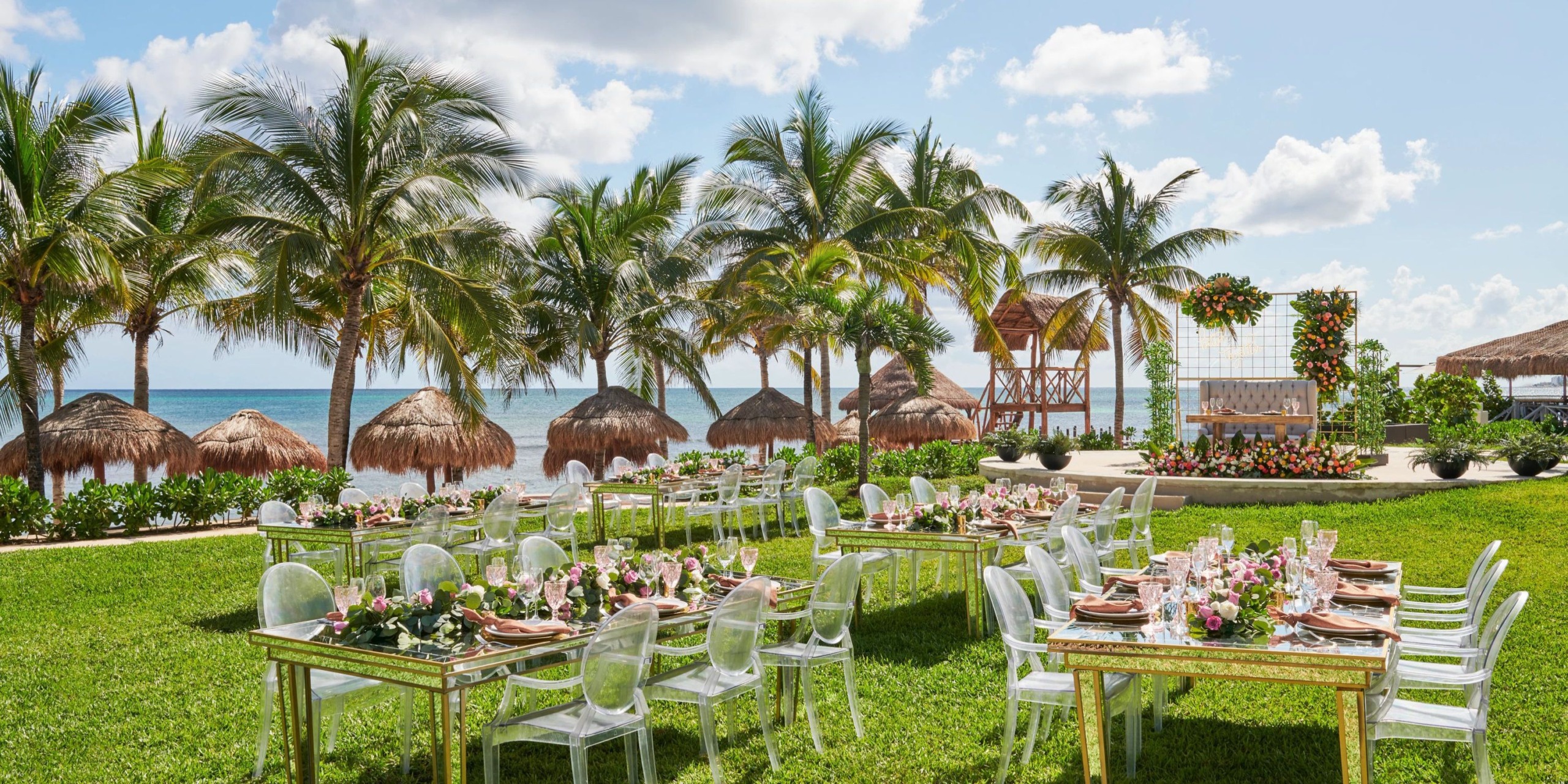 Experience a destination wedding at Hyatt Ziva Riviera Cancun, featuring a picturesque beachside setup complete with lush grass, elegant palm trees, and beautifully decorated tables.
