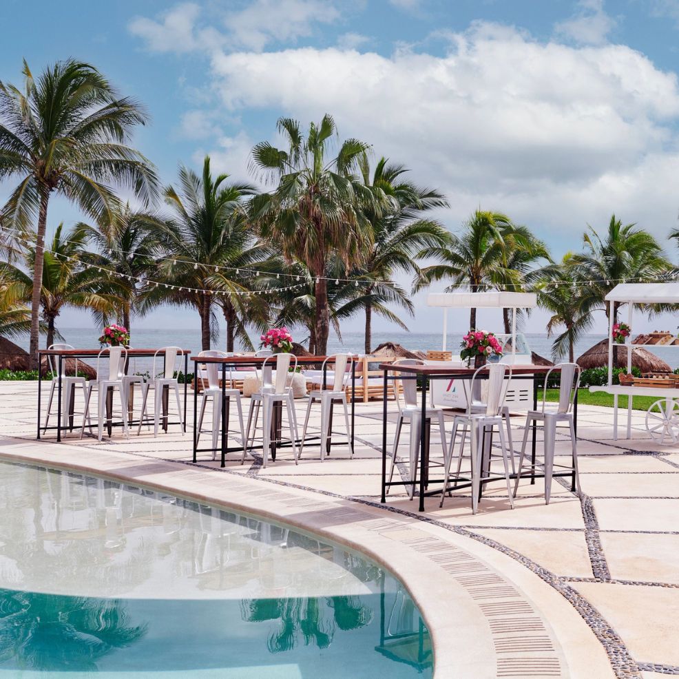 Experience a stunning destination wedding at Hyatt Ziva Riviera Cancun, featuring a picturesque poolside setup adorned with high tables, chairs, and swaying palm trees.