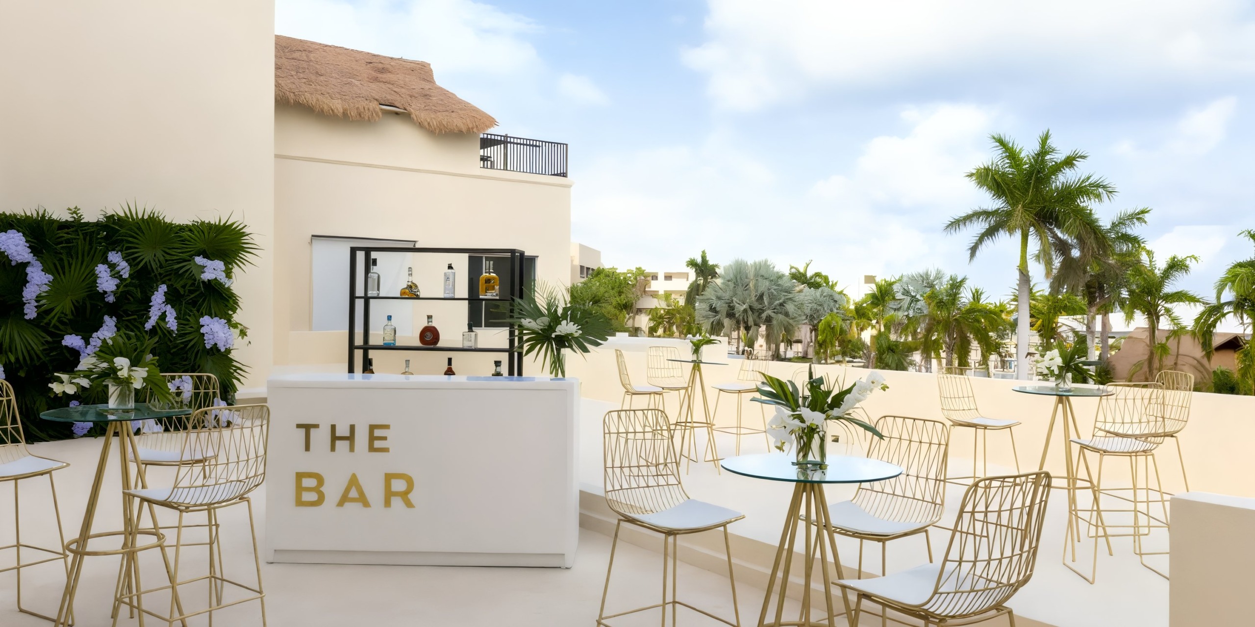 A contemporary outdoor bar at Hyatt Ziva, perfect for a destination wedding, featuring white tables, wire chairs, and surrounded by lush tropical plants.