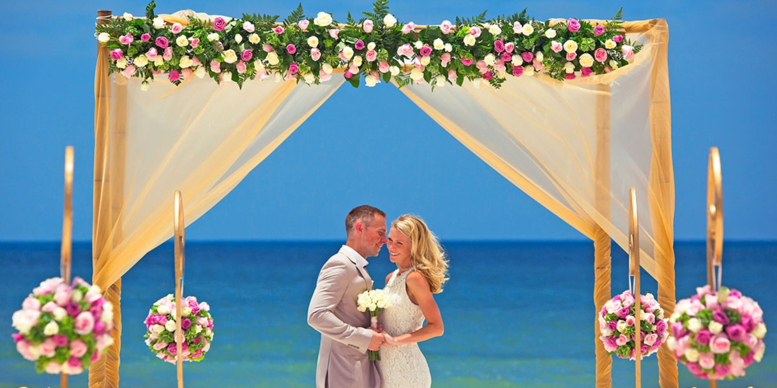 A couple holding hands beneath a floral arch during their destination wedding at Hideaway at Royalton Riviera Cancun beach.