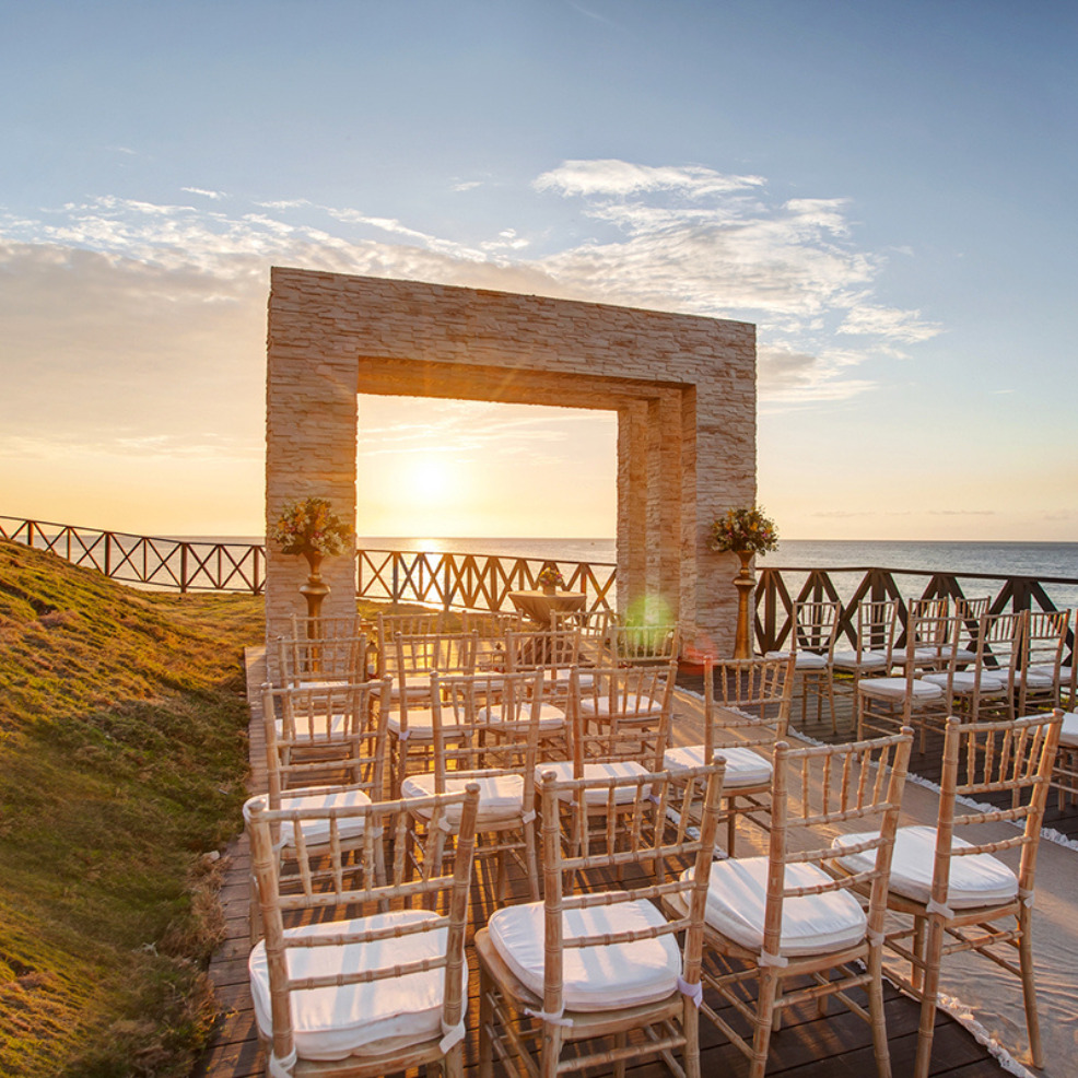 Destination wedding setup with chairs facing a stone arch overlooking the sea at sunset.