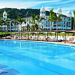 Hotel Riu Palace Costa Rica, with white domed buildings overlooking a large blue swimming pool, perfect for destination weddings, surrounded by loungers under a clear blue sky.