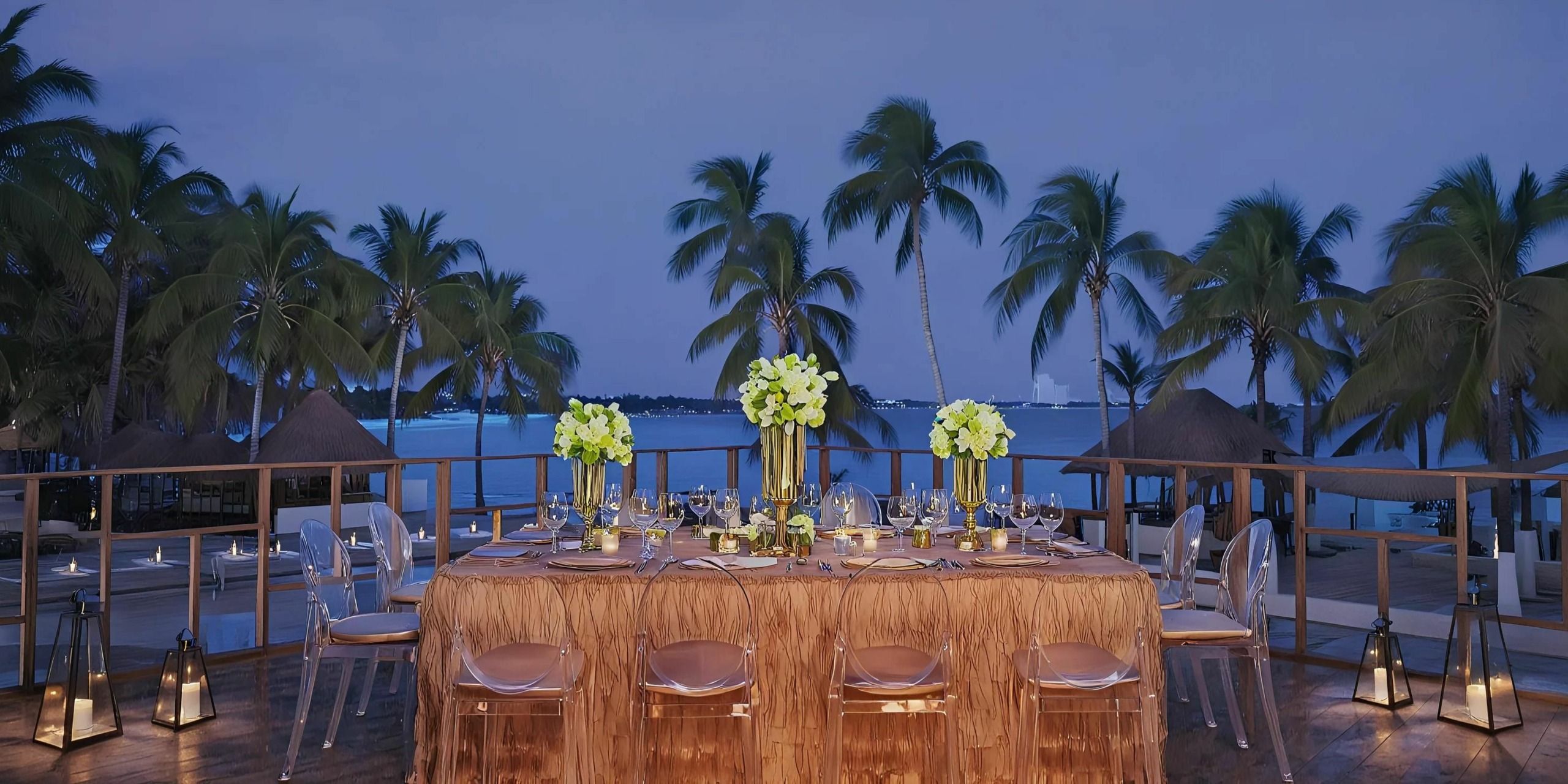 Outdoor dining setup with a large banquet table adorned with green floral centerpieces, set against a backdrop of palm trees and a twilight sky by the ocean.