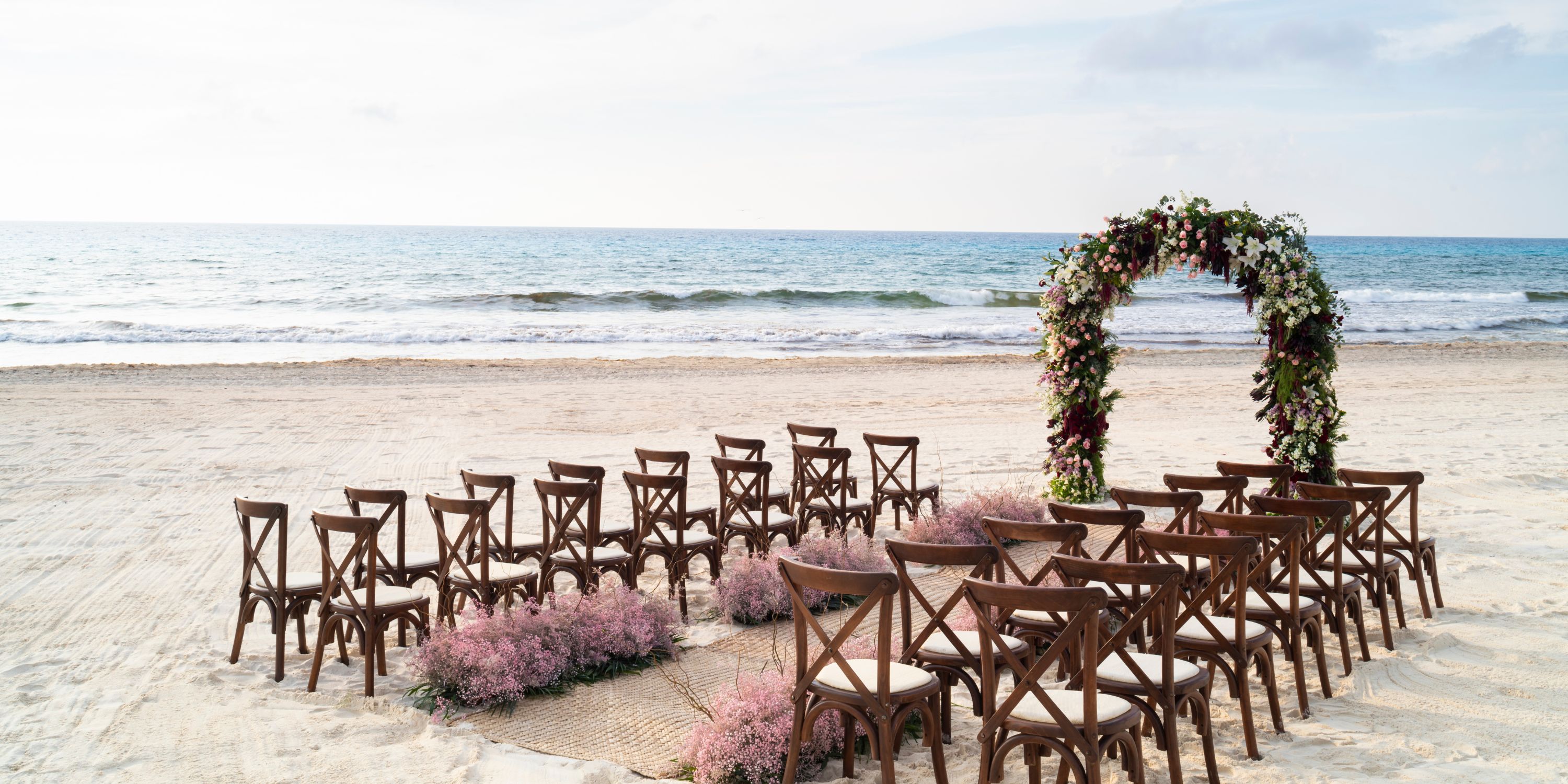 Destination wedding setup with wooden chairs and a floral arch on the sandy beach at Hyatt Zilara Cancun.