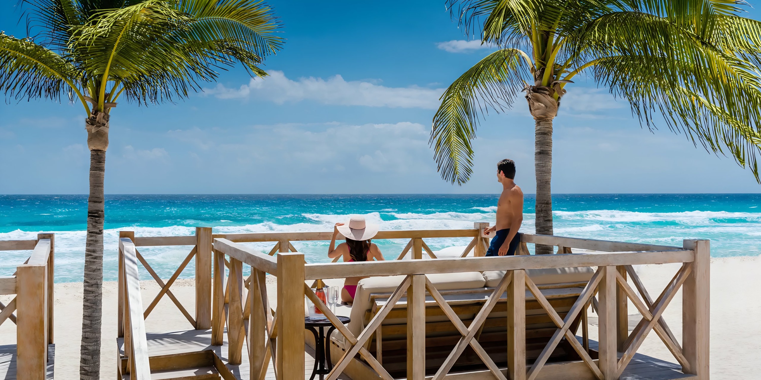 A couple enjoys a sunny beach view from the Hyatt Zilara Cancun, surrounded by palm trees and overlooking vibrant blue ocean waves.