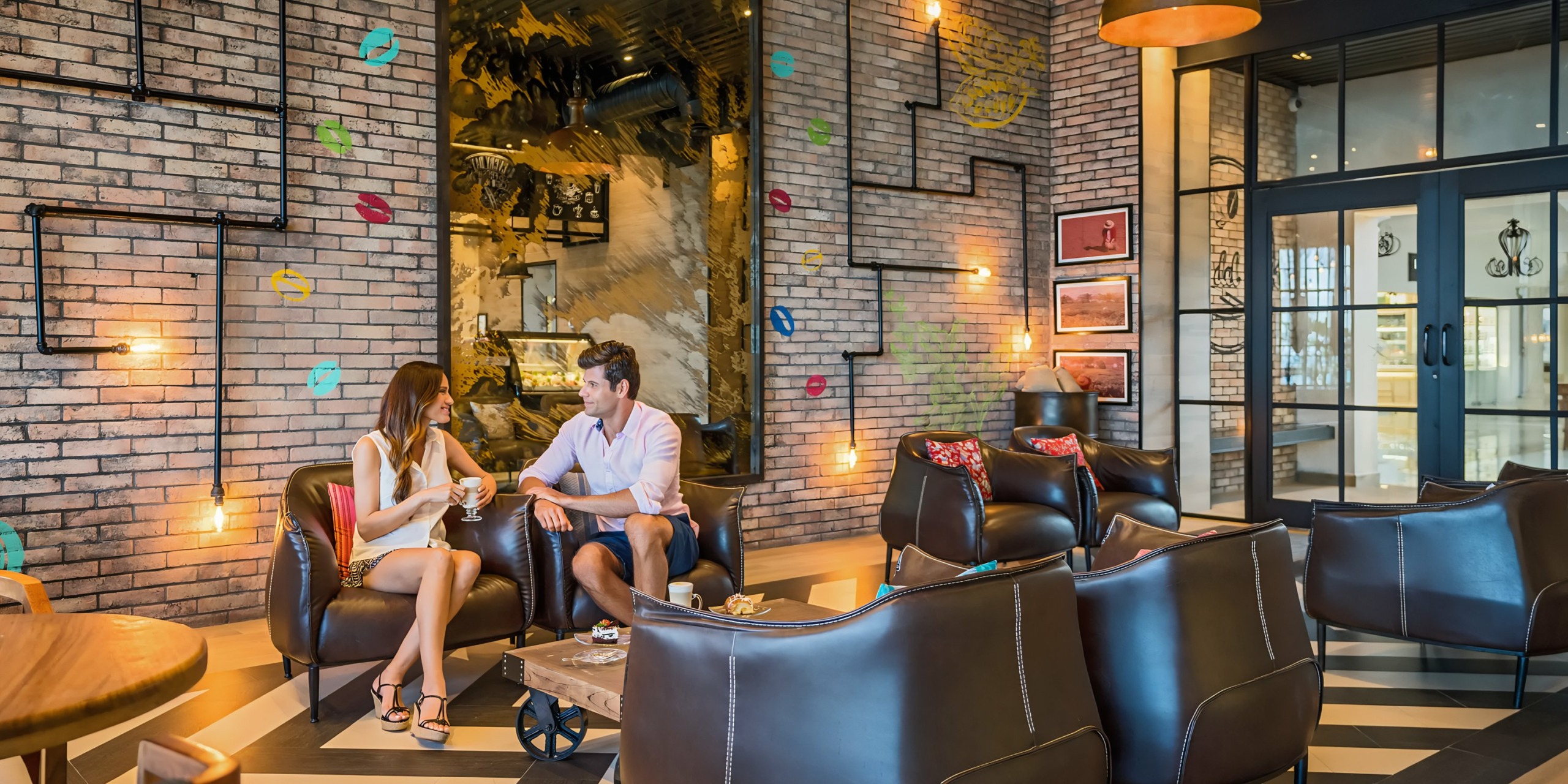 A man and a woman sit chatting in Hyatt Zilara Cancun, a stylish café with brick walls, modern decor, and comfortable seating.