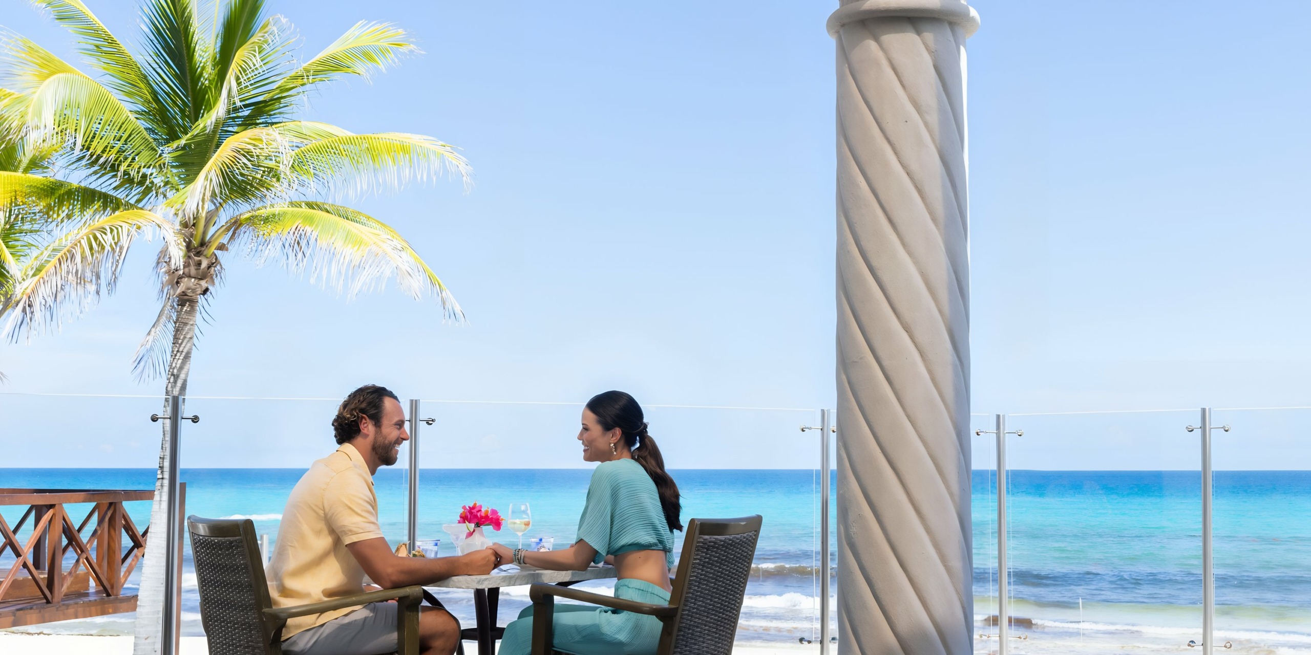 A couple dining at a beachside table under a palm tree, with clear blue skies and ocean in the background at Hyatt Zilara Cancun.
