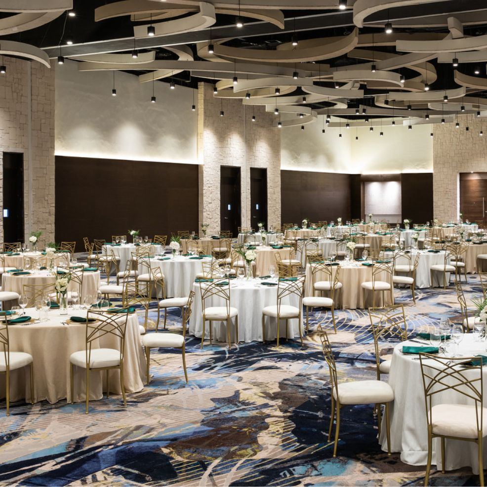A spacious banquet hall, perfect for a destination wedding, features round tables adorned with gold chairs and a beautifully decorative ceiling design.