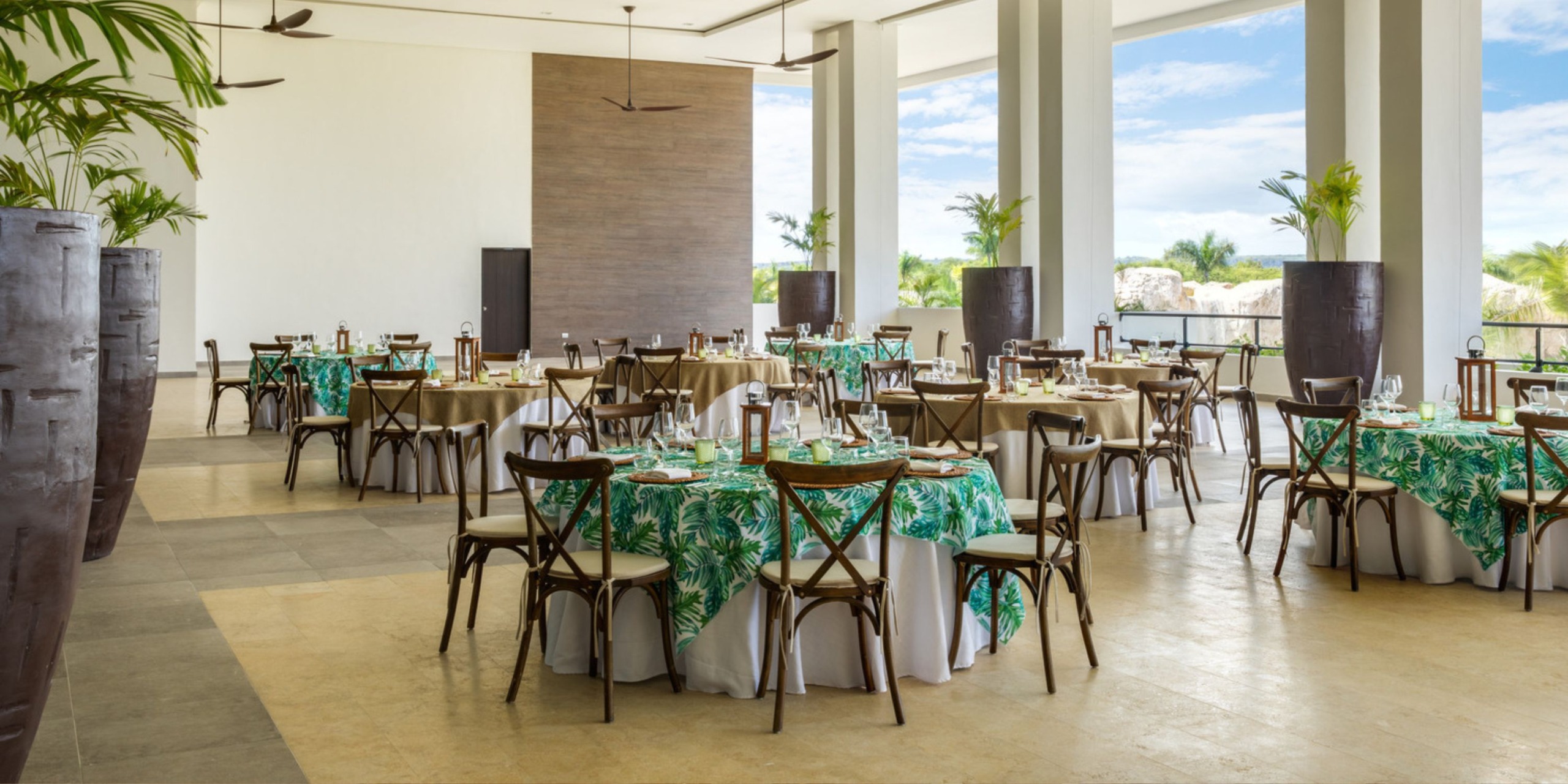 A dining area set for a destination wedding, featuring round tables adorned with green patterned tablecloths and accompanied by wooden chairs.